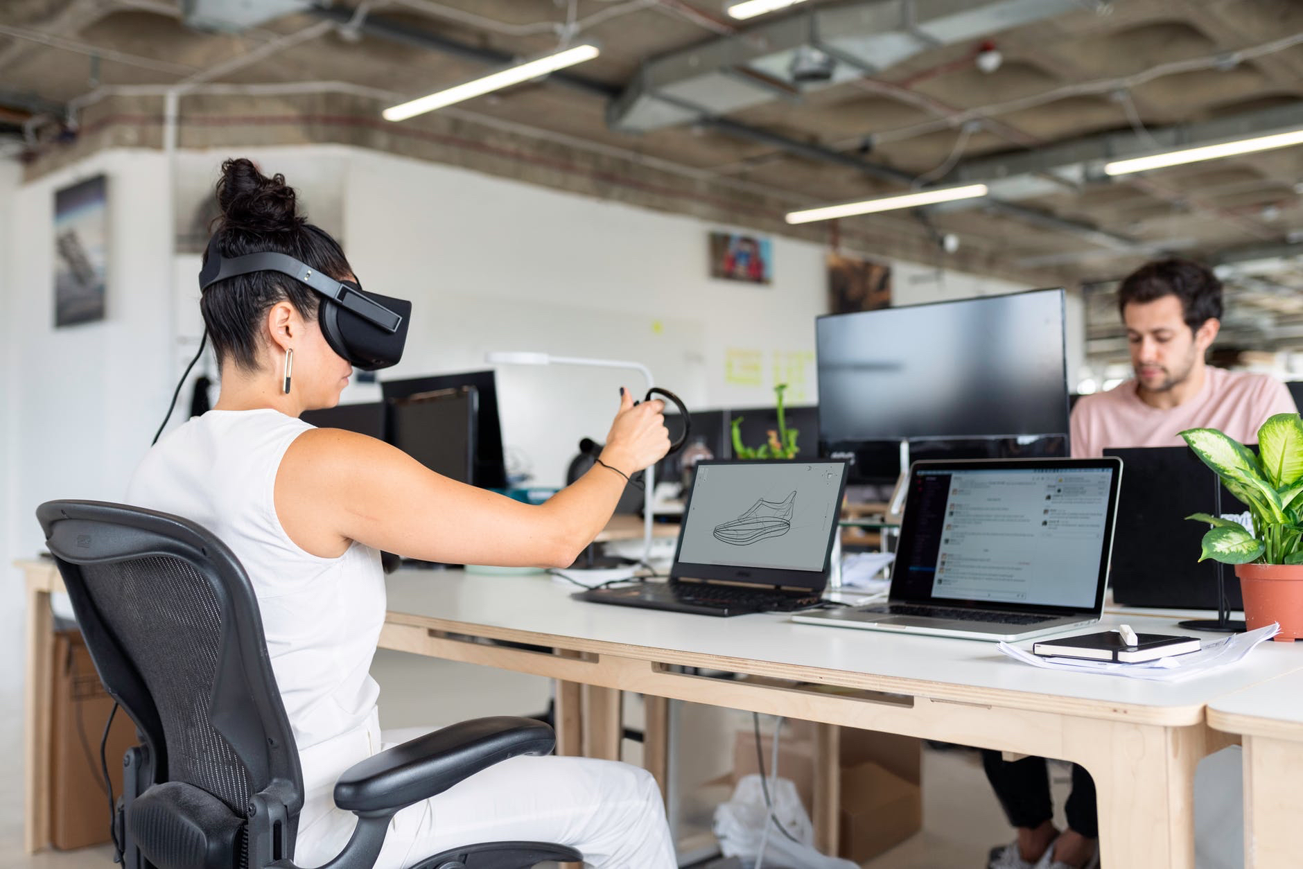 Woman with virtual reality headset seated at desk with computer across from another user without headset.