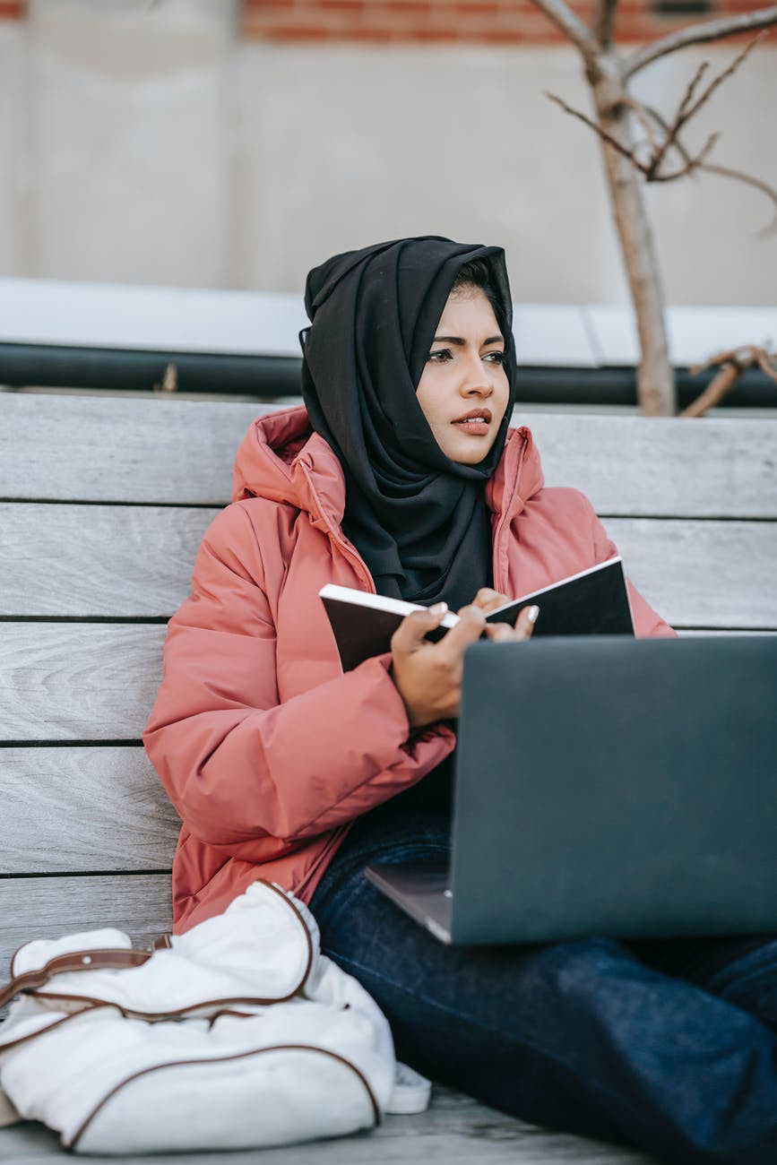 Woman woth head scarf reading book and holding laptop .