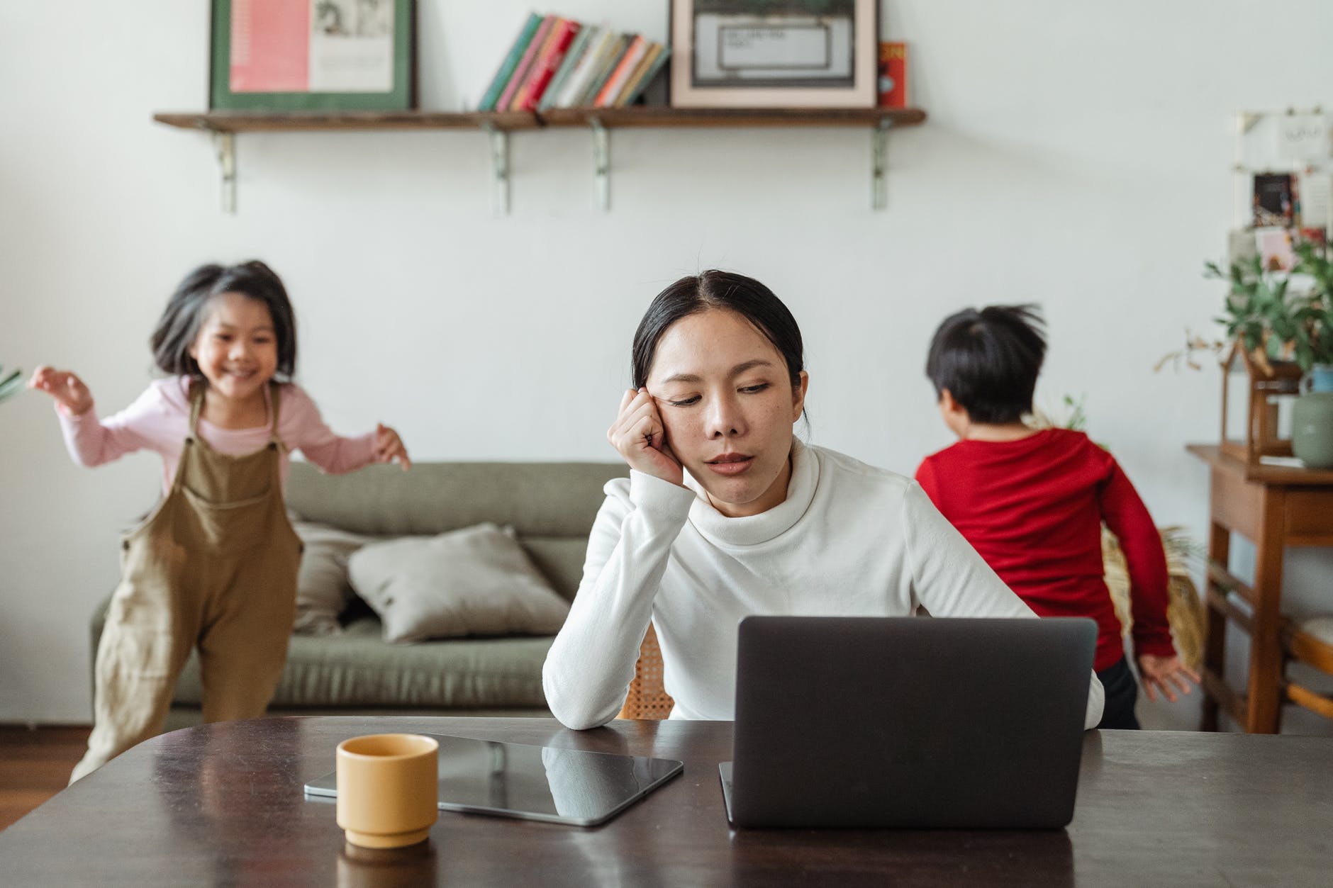 Asian woman working on laptop and two children playing behind. Woman looks tired and is holding face with hand.