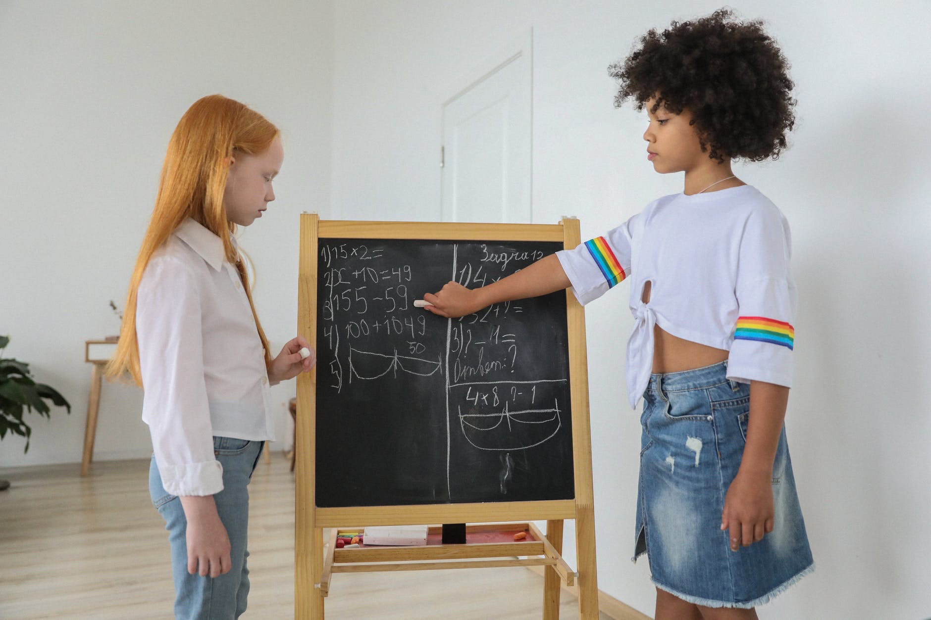Girl with red hair and black girl working together on math problems at small easel chalkboard.