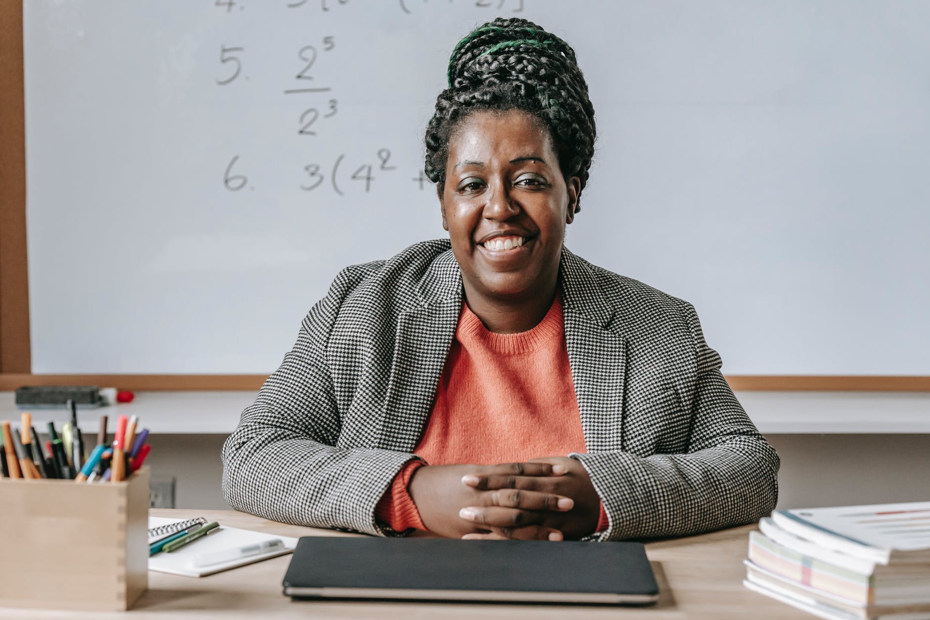 Black female professor seated at desk smiling and whiteboard with math/numbers written behind them.