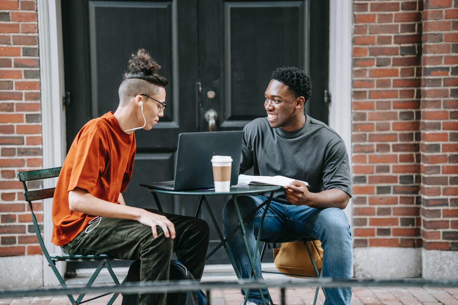 Two men sitting at table outside and working on a laptop. One has a ponytail and is wearing headphones. The other is a black man with a notebook. There is a coffee cup on the small table.