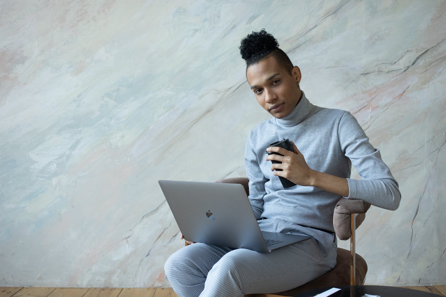 Brown skin male with long hair in bun and holding travel mug while seated at small table with laptop.