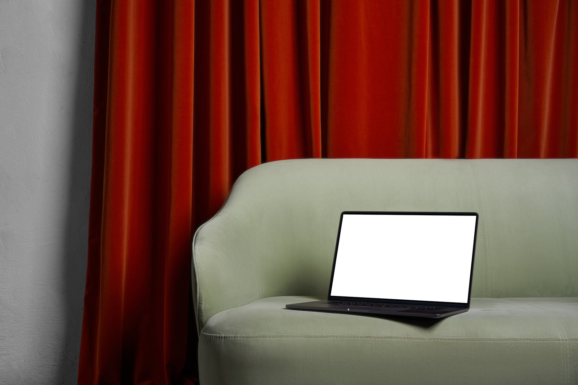 Laptop with white screen on beige couch with with red curtains in the background.
