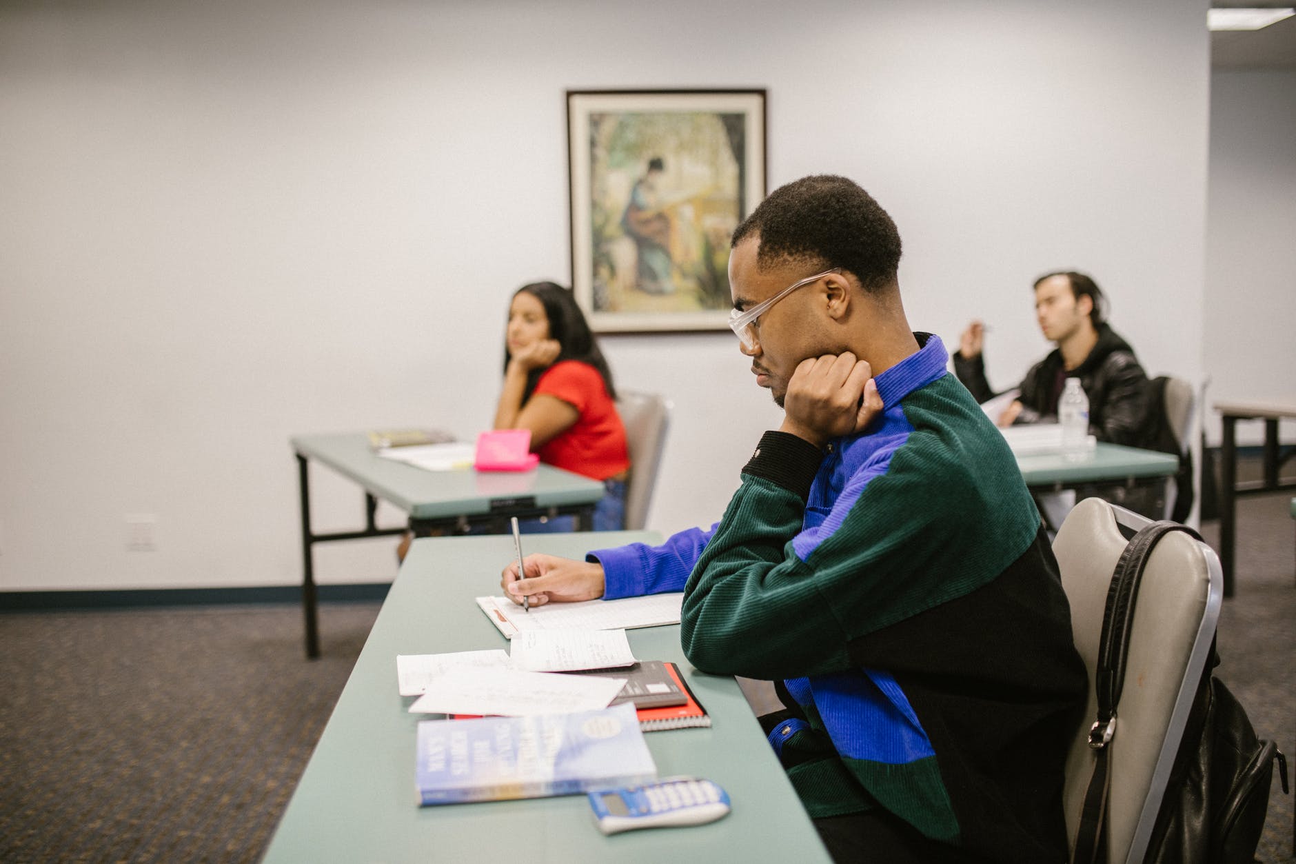 Students at small white desks in class. Black man front center and woman in back left. Person in back right.