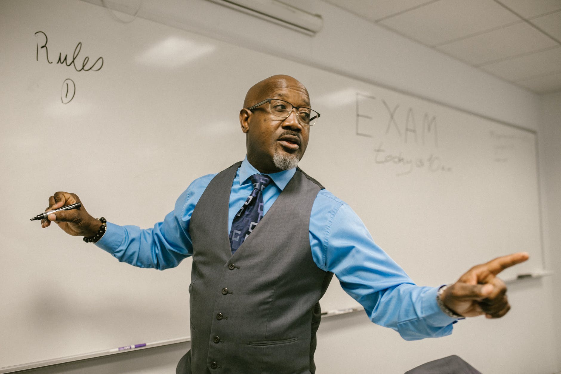 Black male professor responding to questions in front of whiteboard. The word "Exam" is written on the whiteboard.