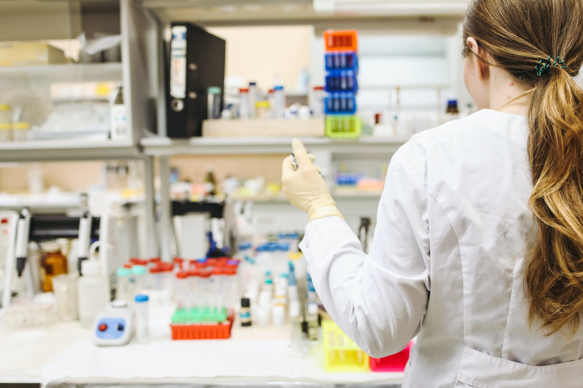 Back of a scientist wearing a white lab coat with long hair in a ponytail facing laboratory bench.