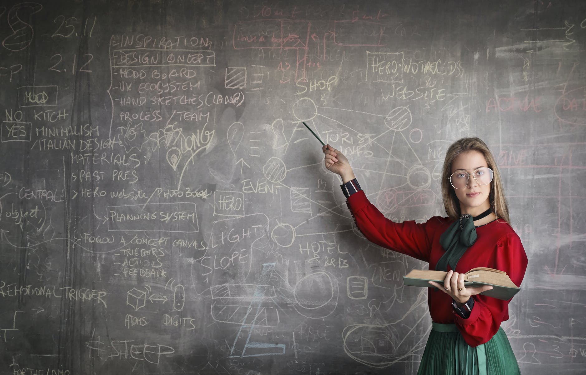 Girl with blond hair an red sweater at chalkboard pointing with pencil.