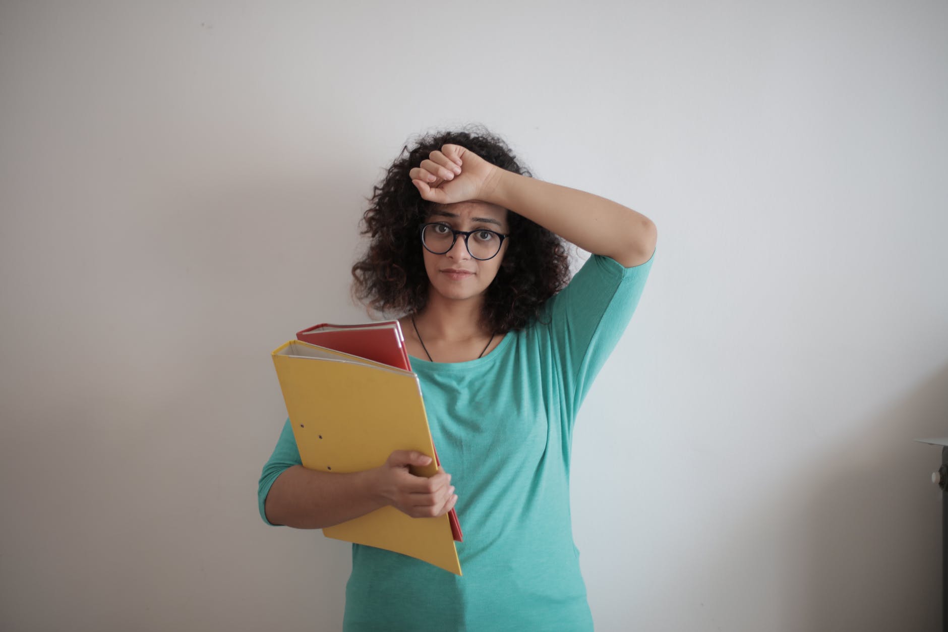 Woman with log curly hair, brown skin, and black glasses with hand on forehead and holding binders.