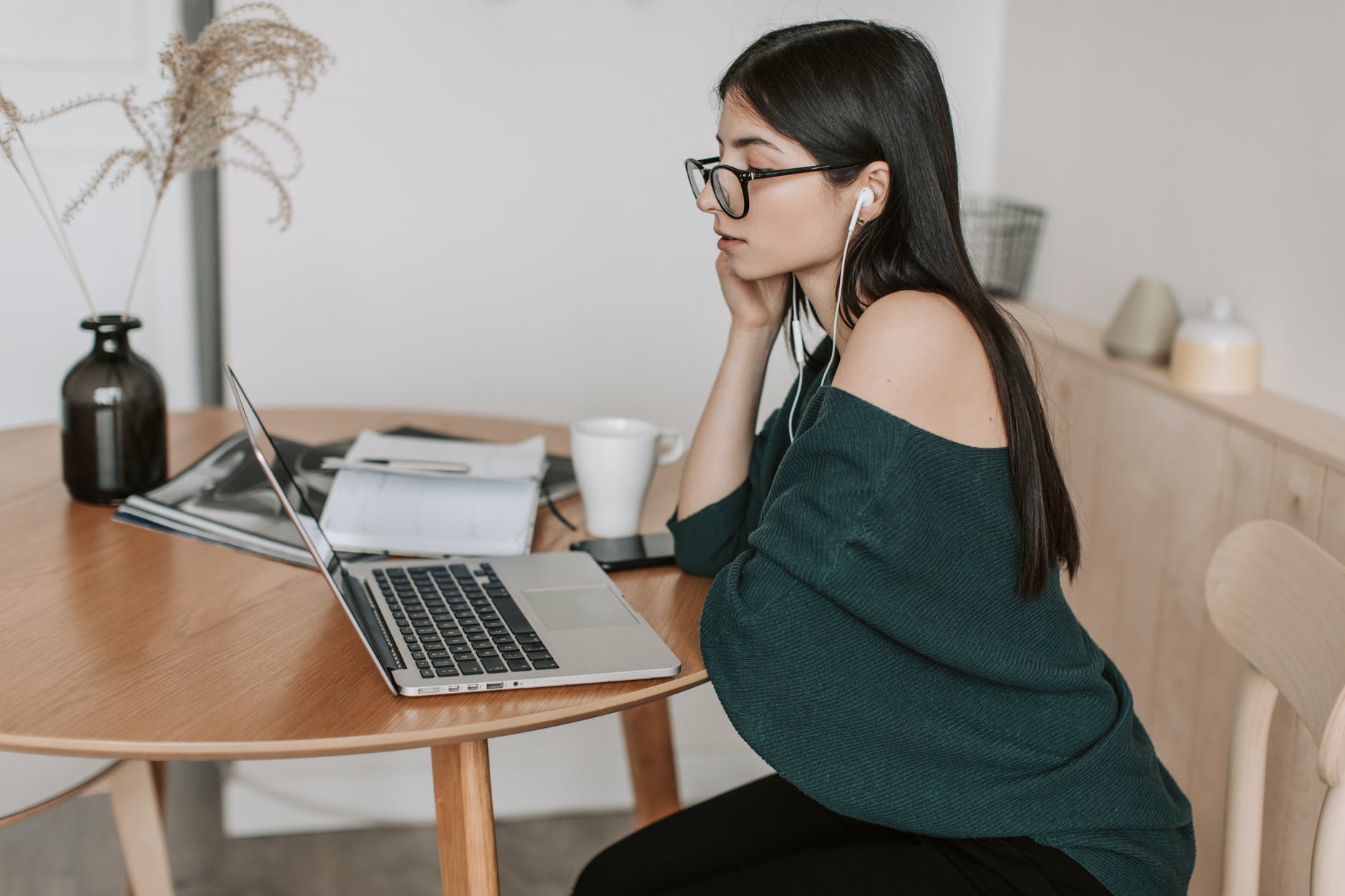 Woman with long black hair seated at table and working on laptop while wearing earbuds.
