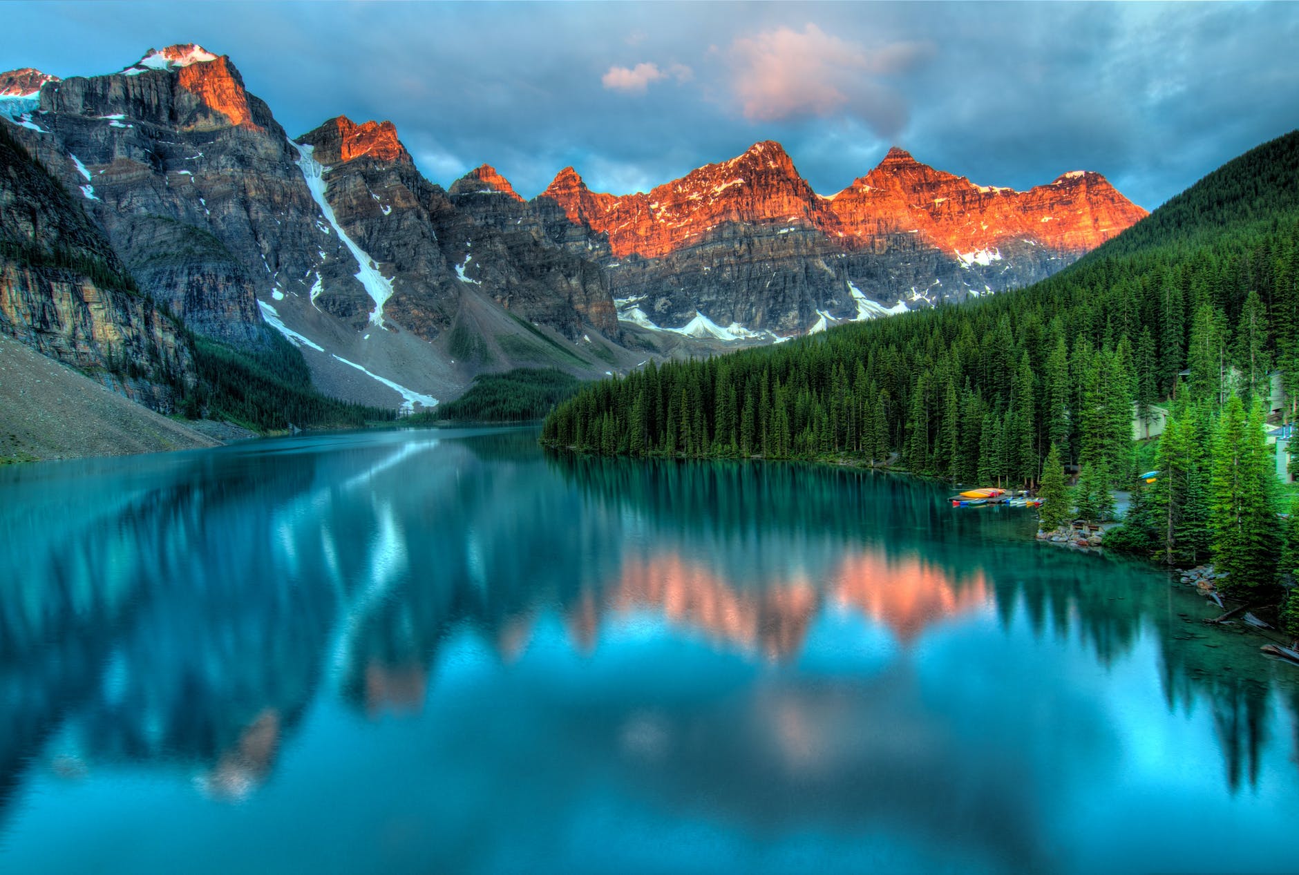 Mountains and clouds in the background. Water reflecting the mountains with snow. Trees on the right and a yellow kayak.