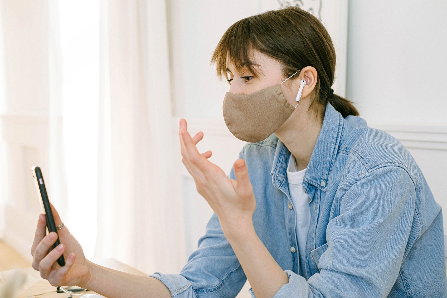 Woman wearing brown face mask and holding phone in front of face with one hand and other hand is gesturing.