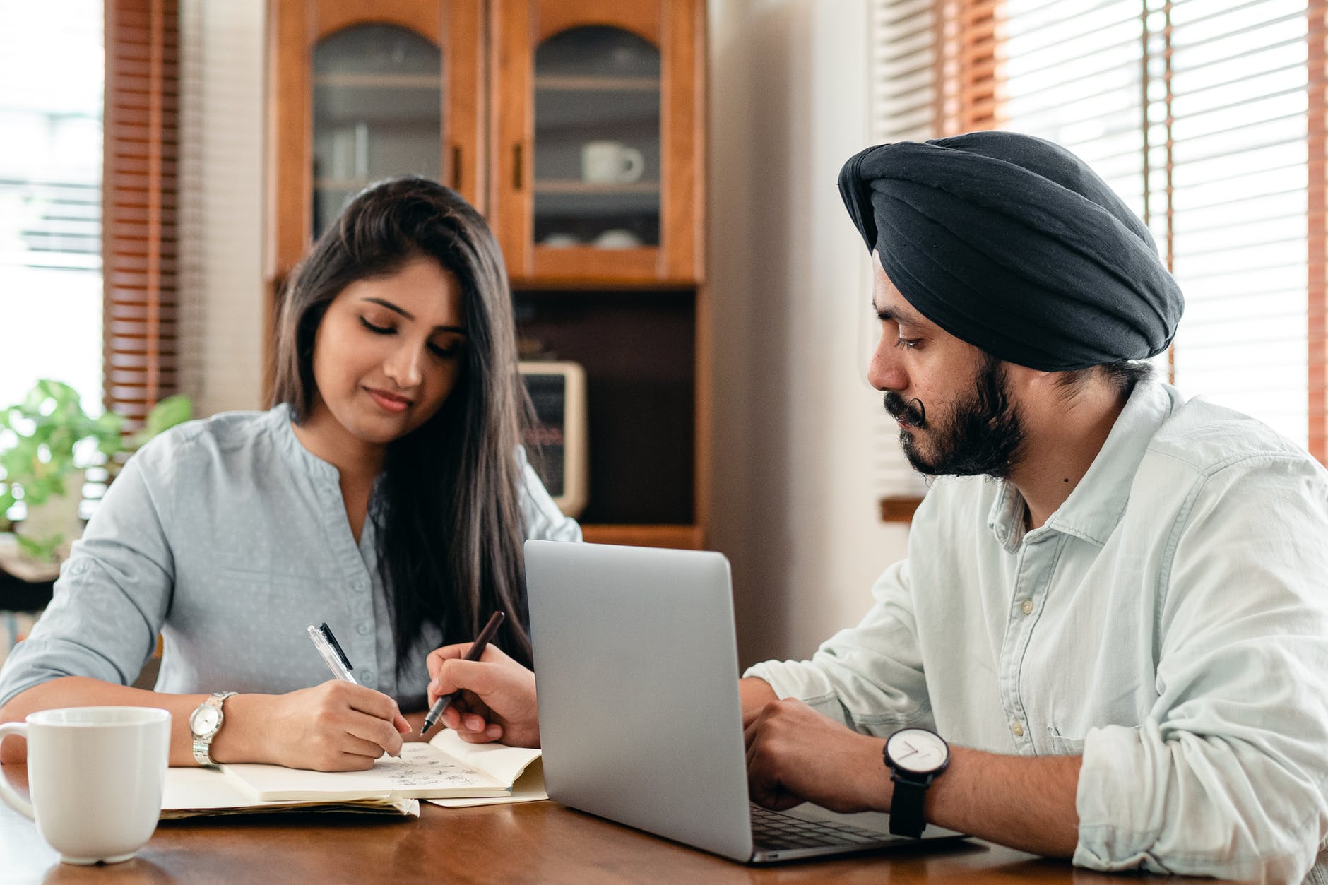 Man and woman working at table with one laptop open. Man has mustache and is wearing a black head covering and is pointing to paper with pen.