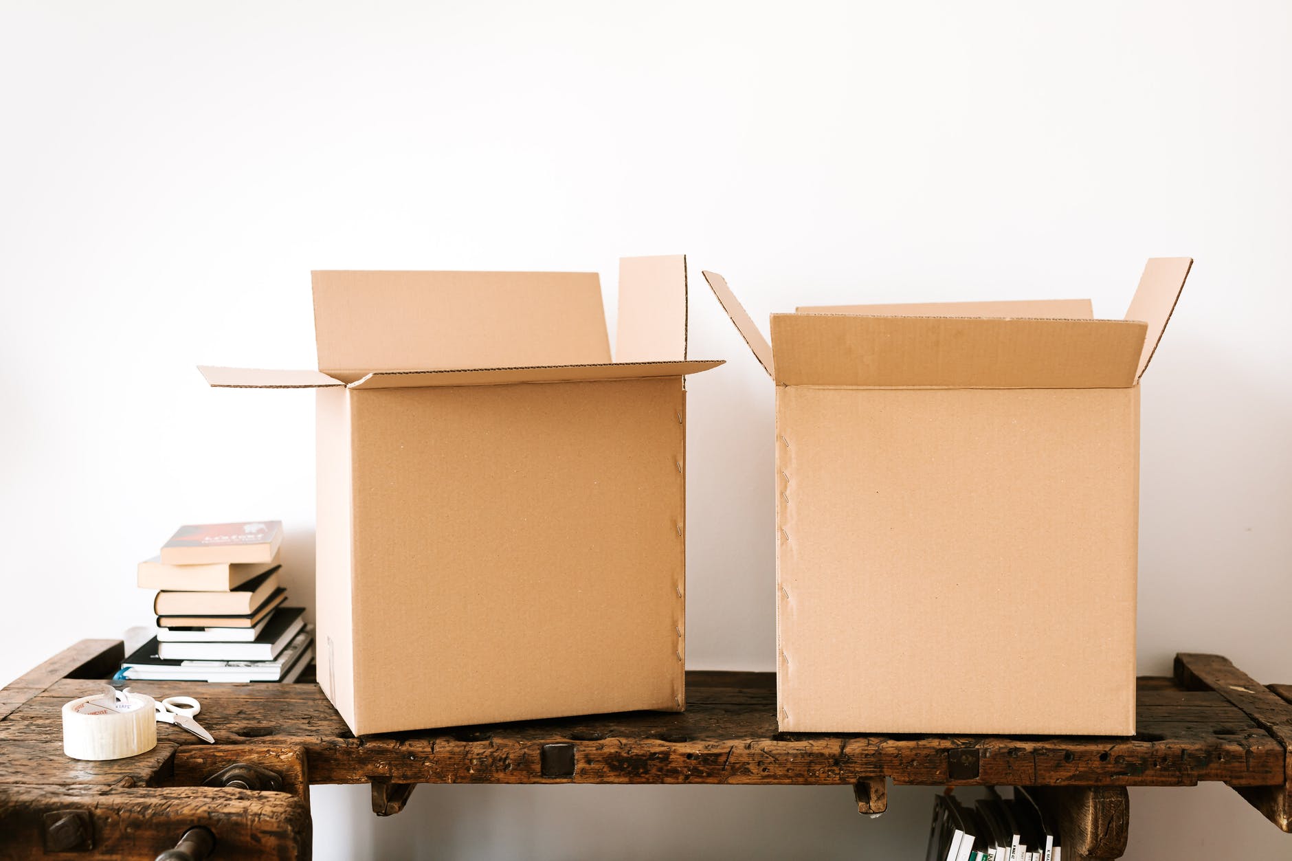 Two cardboard boxes on a wooden bench. The boxes are open. Packing tape and scissors are on the left of bench. Books are placed behind the box on the left.