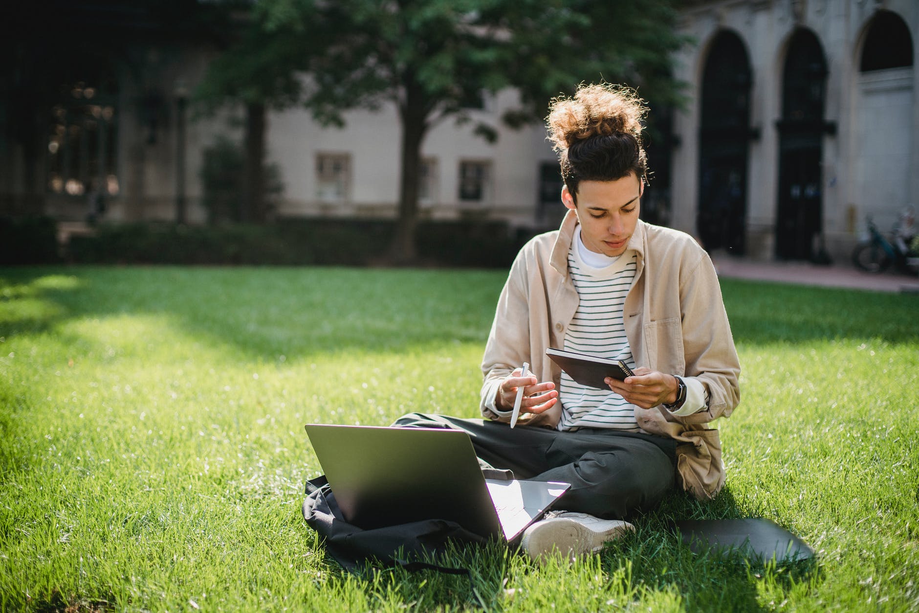 Man with curly black hair in bun sitting on grass with laptop and notebook.