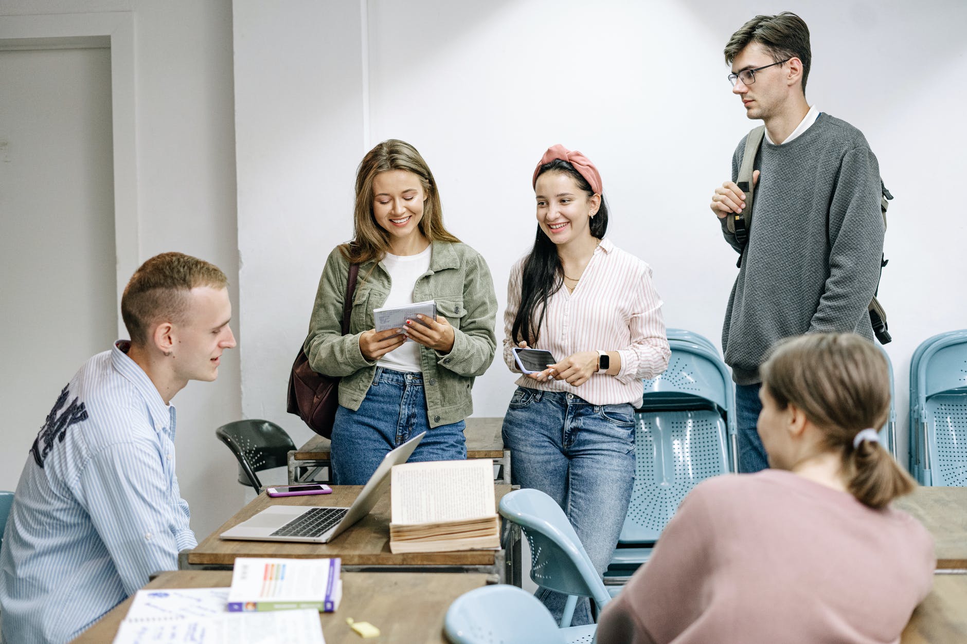 Five high school students working around a table with a laptop. From left to right: white boy seated at desk with short hair, two teenage girls standing, boy watching and discussing with the group, and the back of another student.