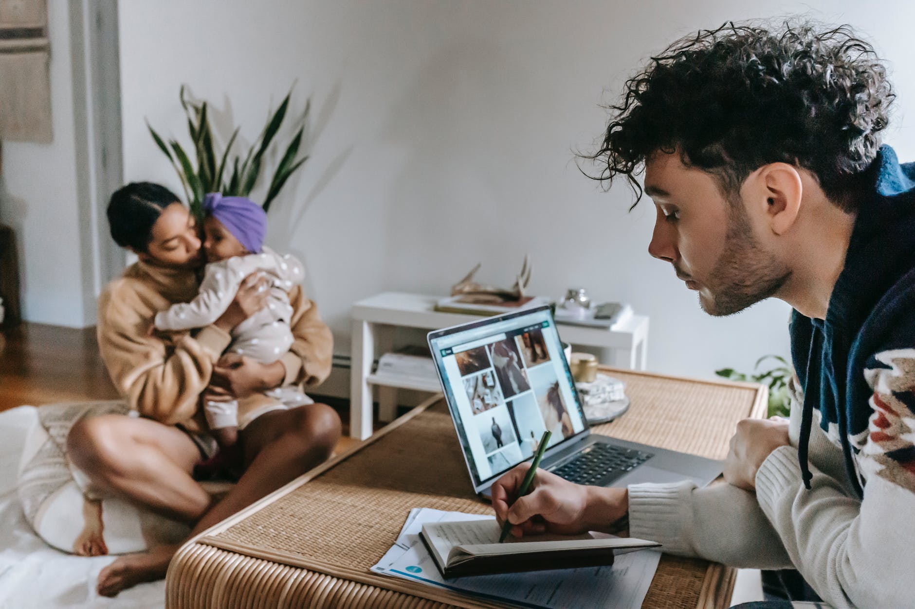 Woman holding baby and man working on laptop with photos on screen.