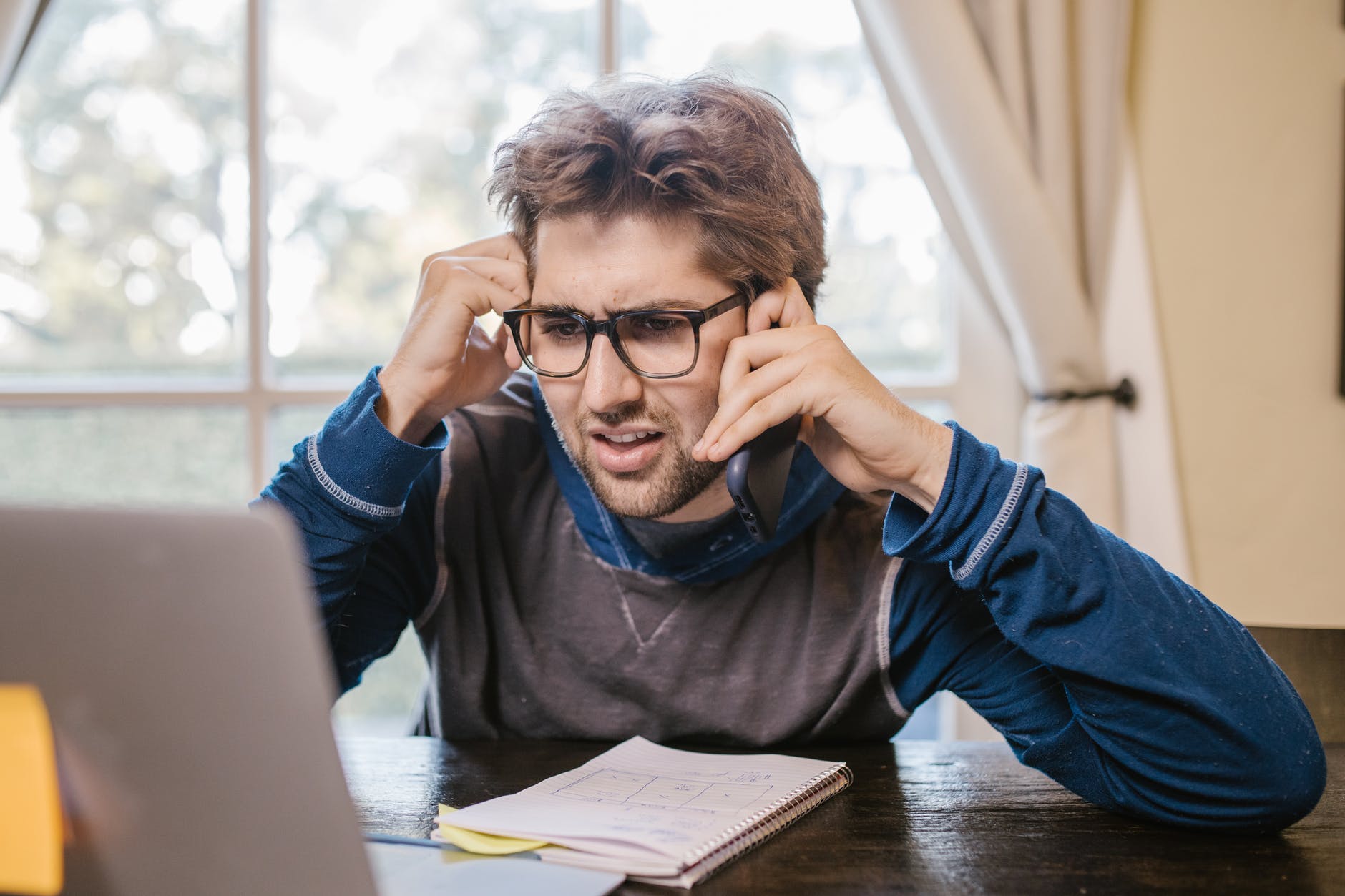Man in front of laptop holding cell phone near ear. Man is wearing glasses and has a notebook on the desk.