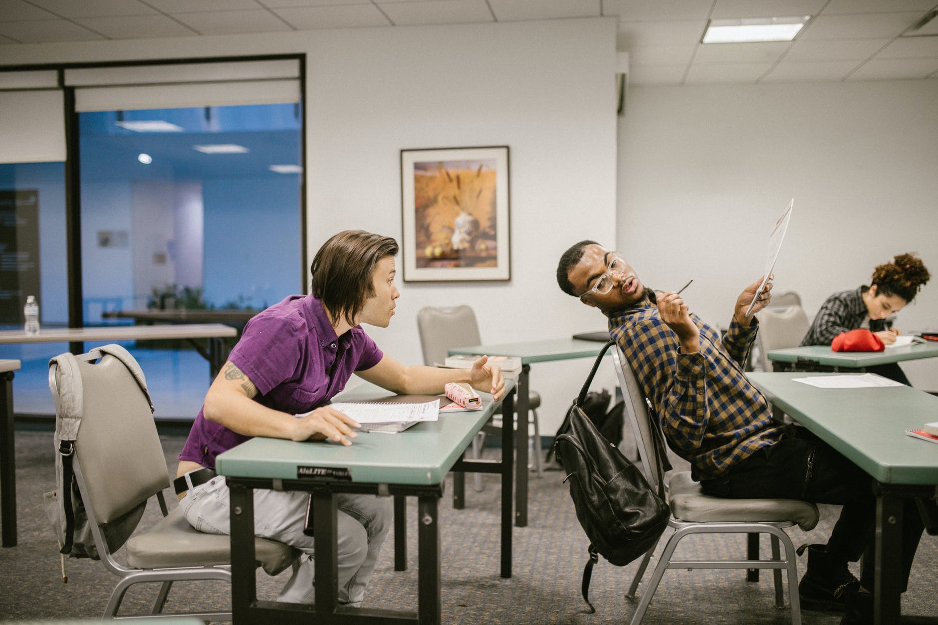 Students in classroom with white tables talking to each other. One black man leaning back and speaking to another person behind them.