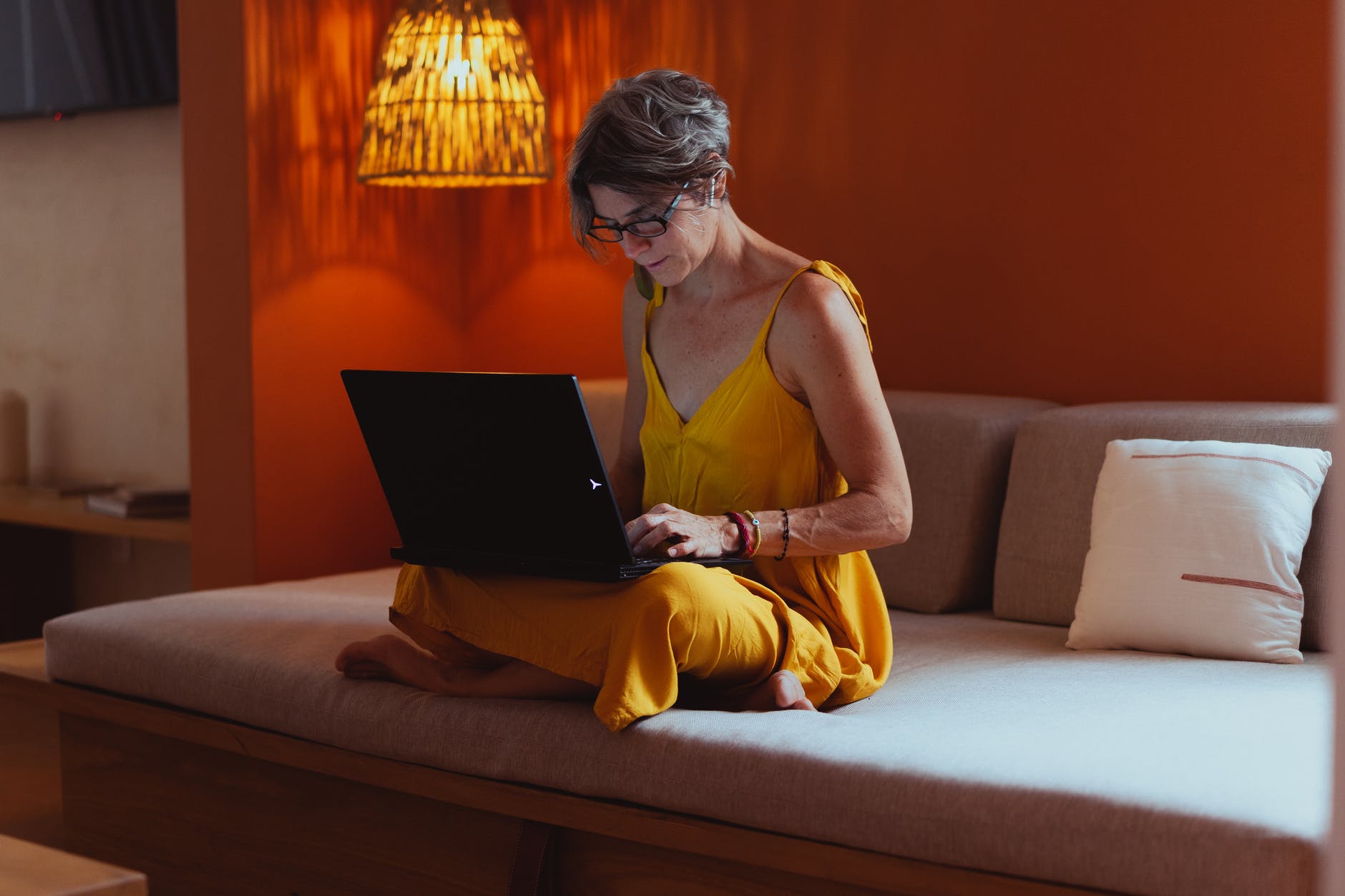 Woman wearing yellow dress sitting on couch working on black laptop. The woman has short brown hair and is seated on a beige couch. The wall behind is dark orange.