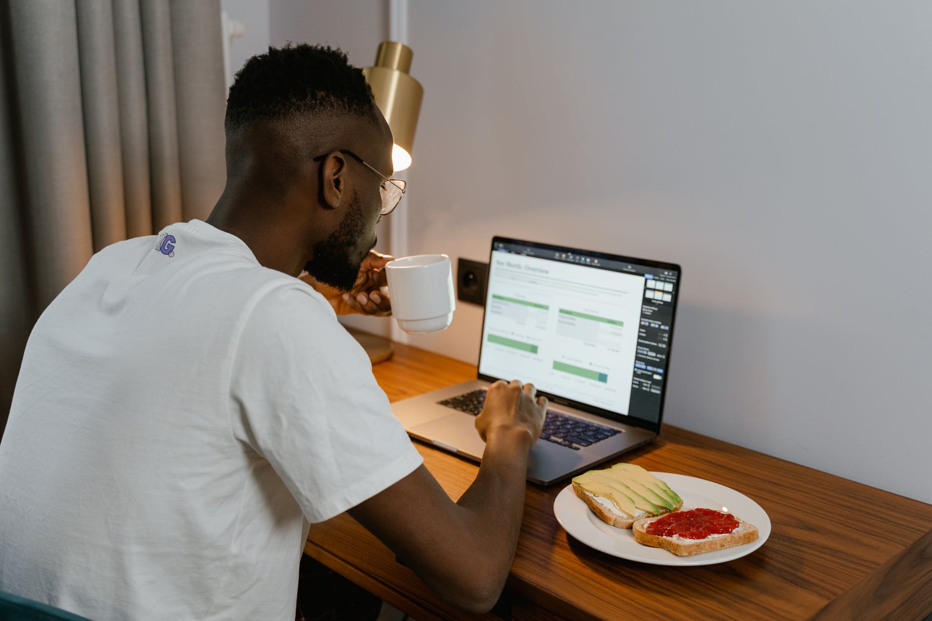 Black man with short hair and beard wearing white t-shirt and holding white tea cup while typing on laptop at wooden desk. Desk has two slices of bread: one with avocado slices and one with red spread.