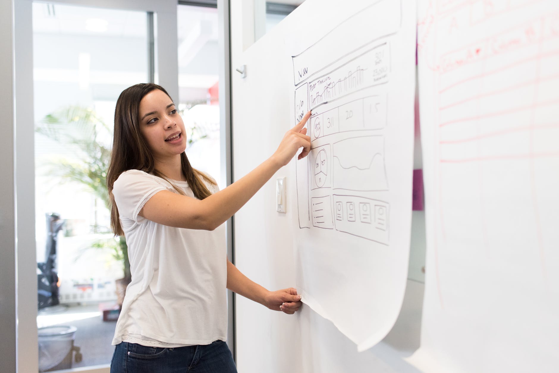 Woman with white t-shirt and long brown hair pointing to paper with graphs on wall.