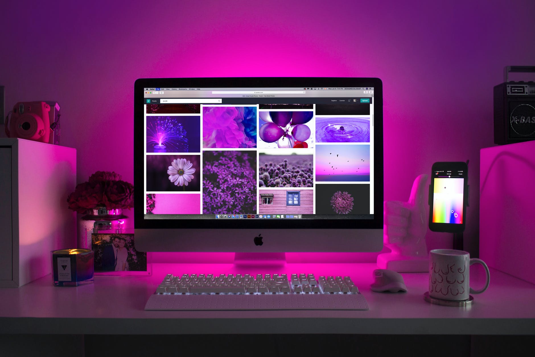 Purple light on wall and desktop, keyboard and cell phone on desk. The screen on the desktop has purple photographs of flowers, water, and the sky. The cell phone to the right is displaying colors. To the left, there is a candle.
