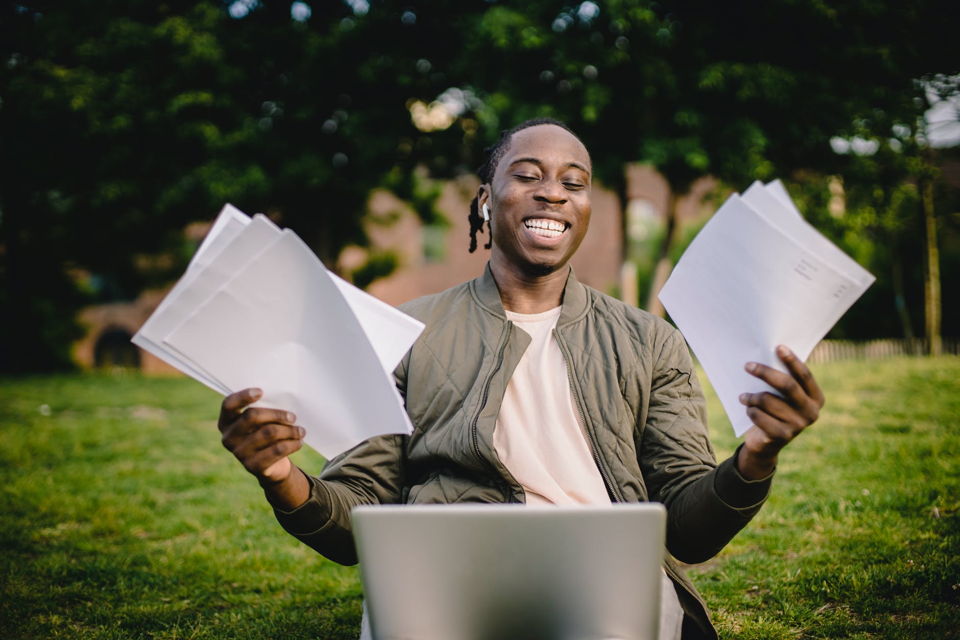 Black man smiling while holding white papers in both hands in front of a laptop.