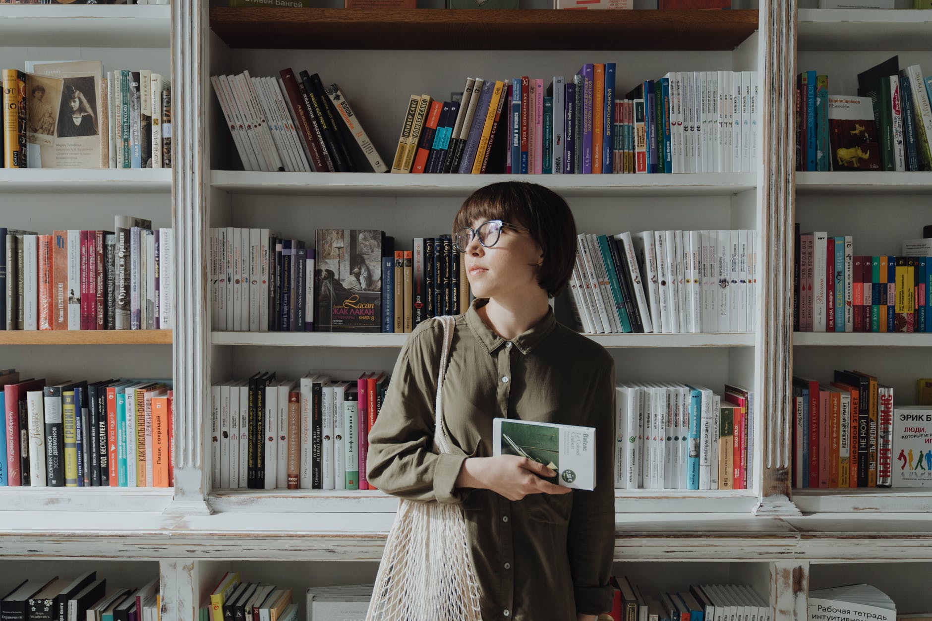 Person with glasses holding book in front of bookcase with numerous books.