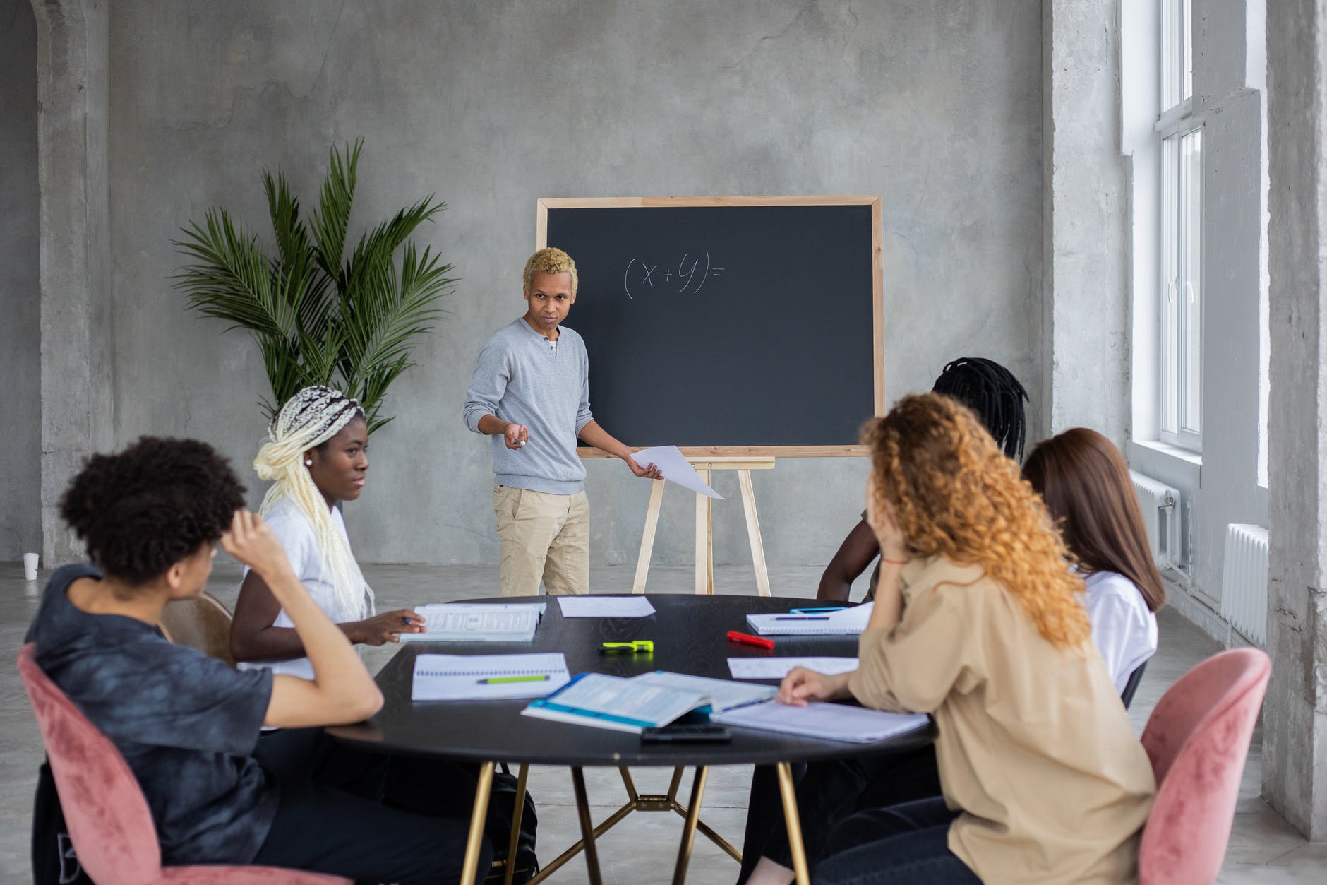 Black man with blond curly short hair writing on chalkboard (x+y)= in front of a table with five people including a woman with a head scarf. The table has several notebooks and highlighters.