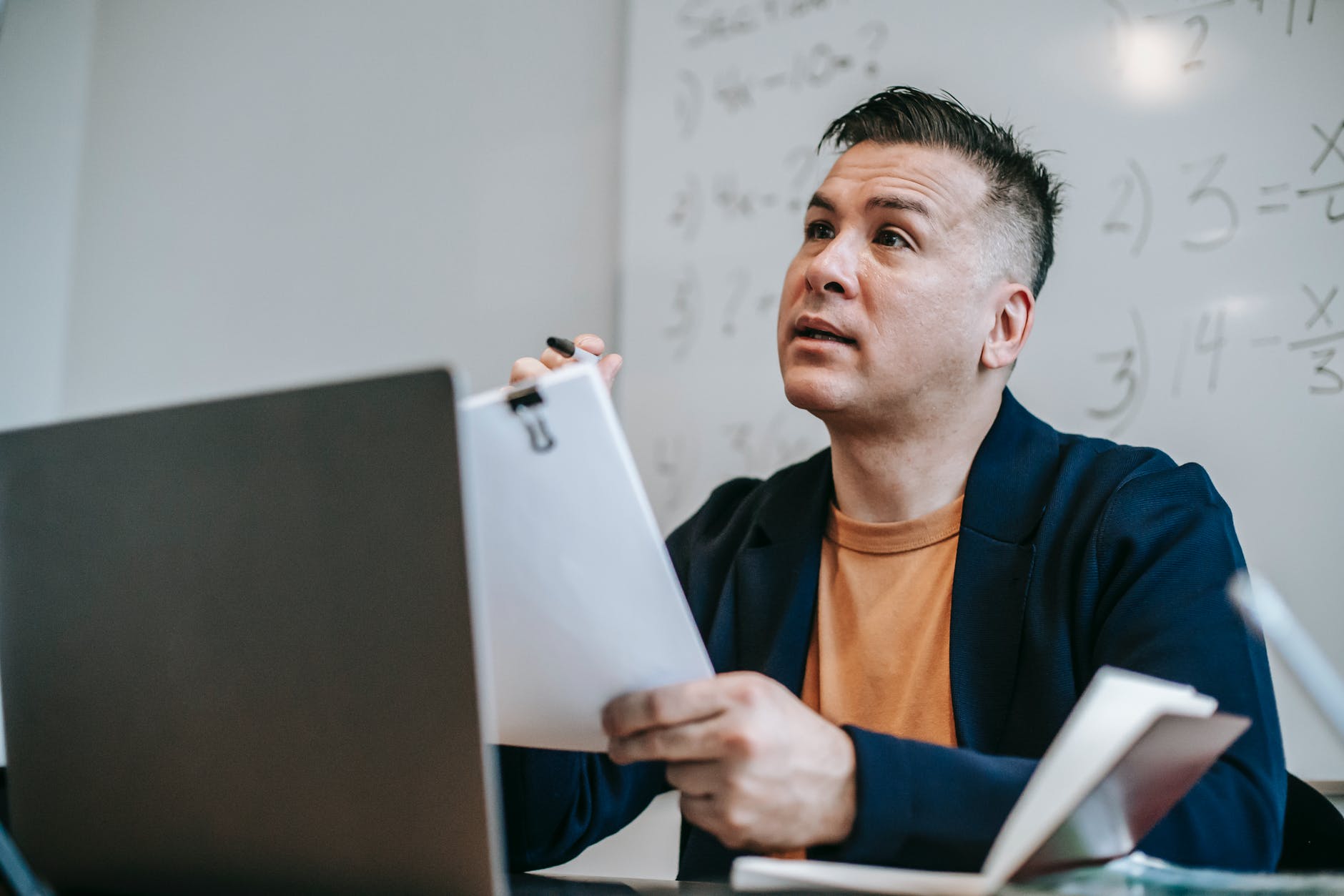 Man with short hair holding papers with binder clip with one hand and marker with another in front of what appears to be a desktop computer monitor.