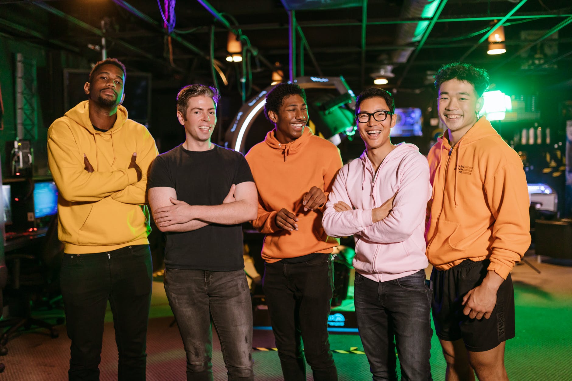Group of men standing up in bar. Left to right, tall black man with yellow hoodie, white man with black t-shirt, black man with orange hoodie, Asian man with white hoodie and glasses, Asian man with orange hoodie.