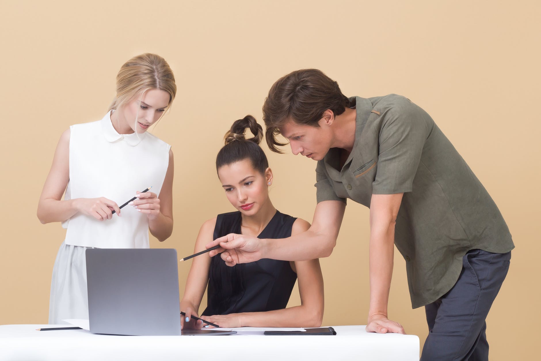 three people looking at laptop. Blonde woman standing, brunette sitting, and man standing leaning on table pointing at laptop with pencil.