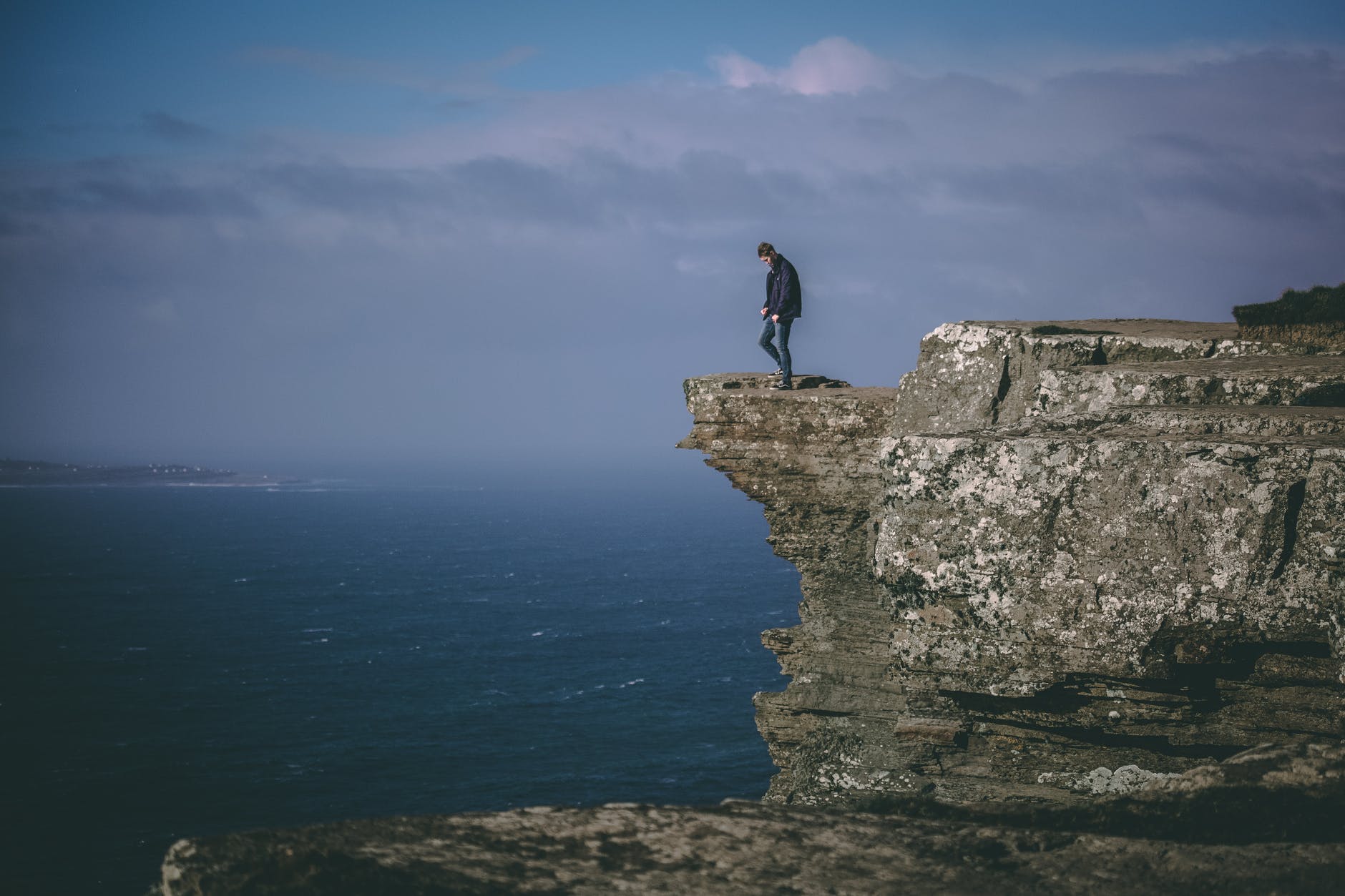 Cliff and person looking down at waves. Cloudy skies are visible.