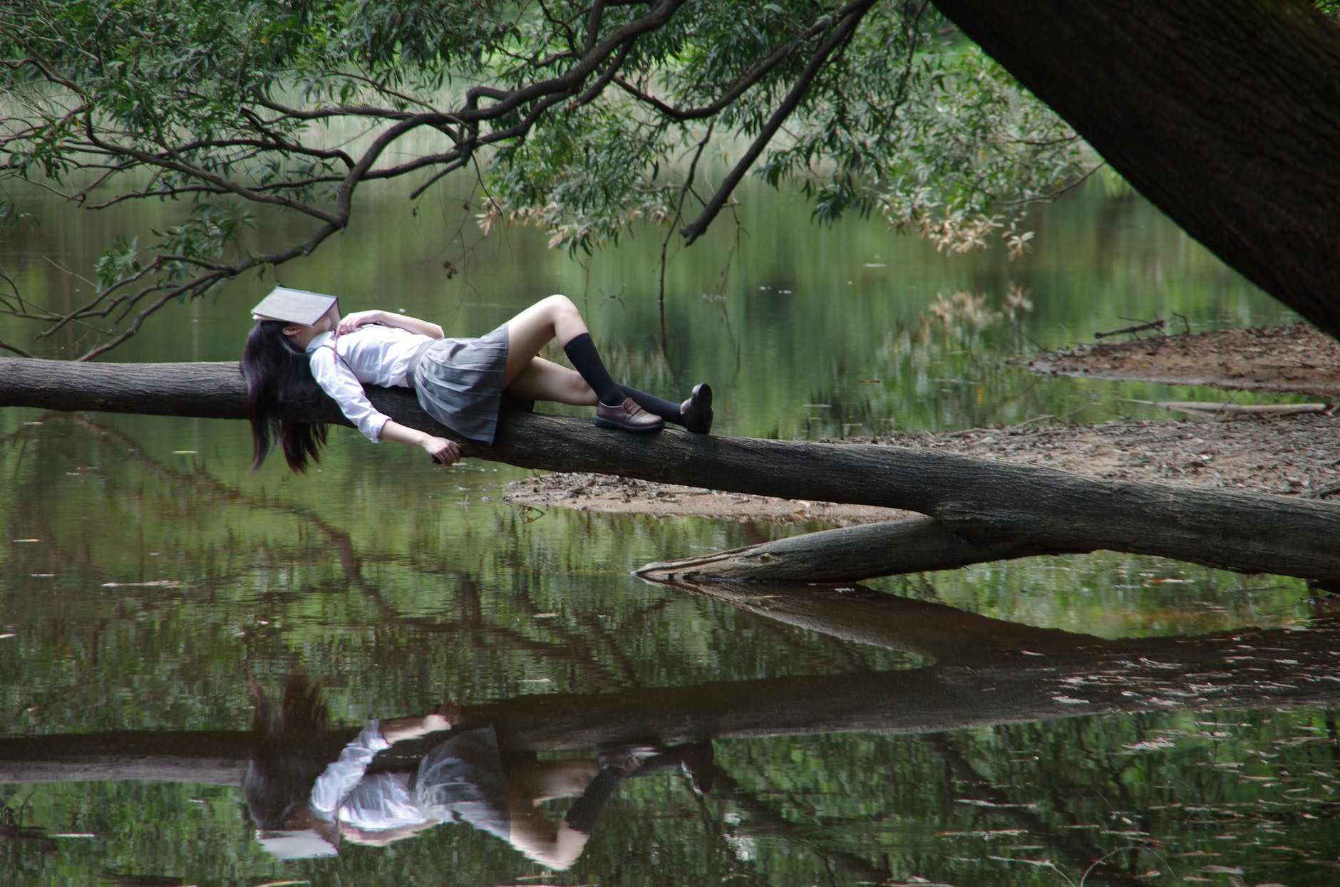 Woman with skirt and long hair on log over water. Woman has book on face. The reflection of person is visible in the water.