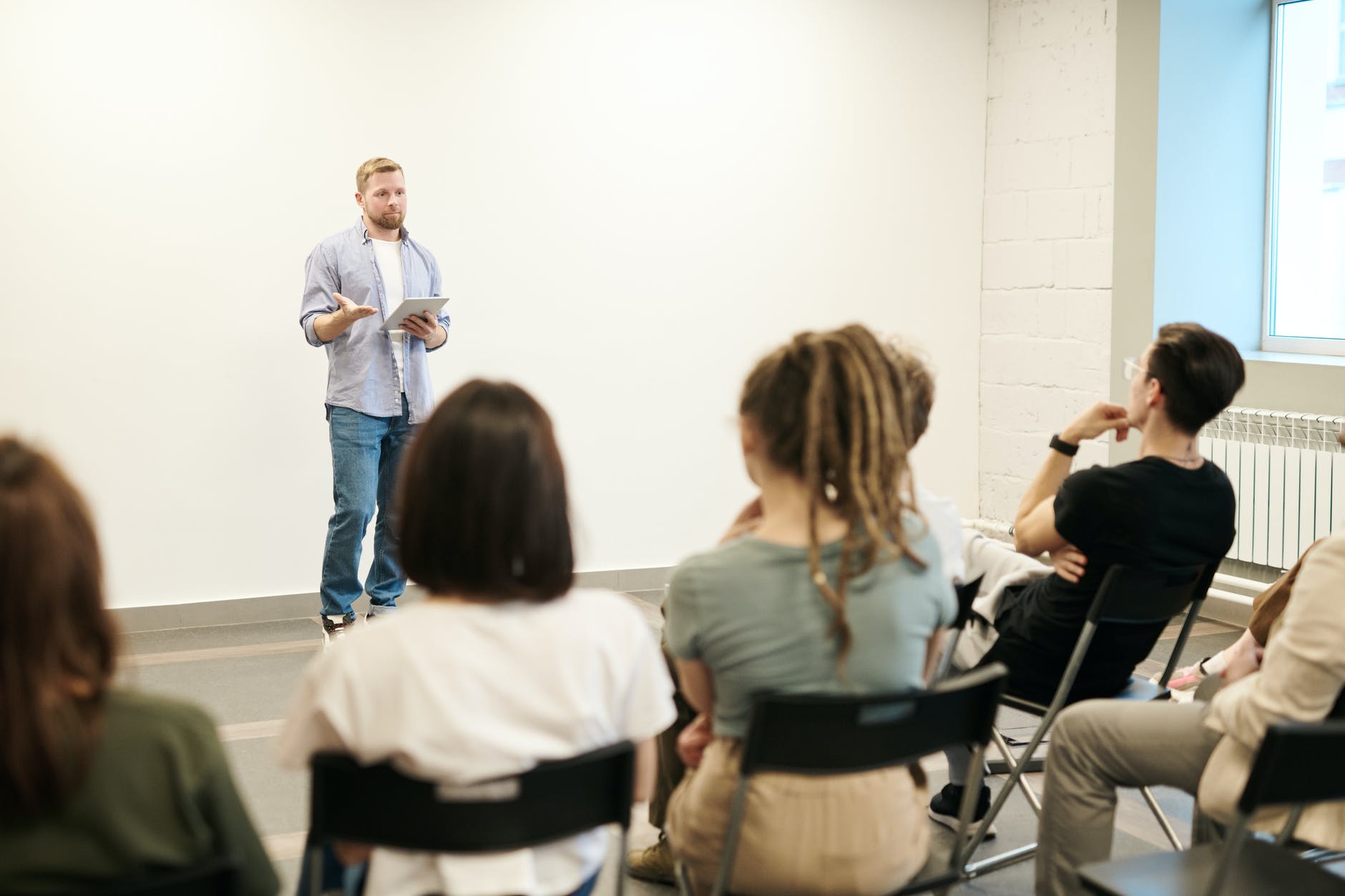 Man presenting while holding tablet. Students are seating in chair facing man.