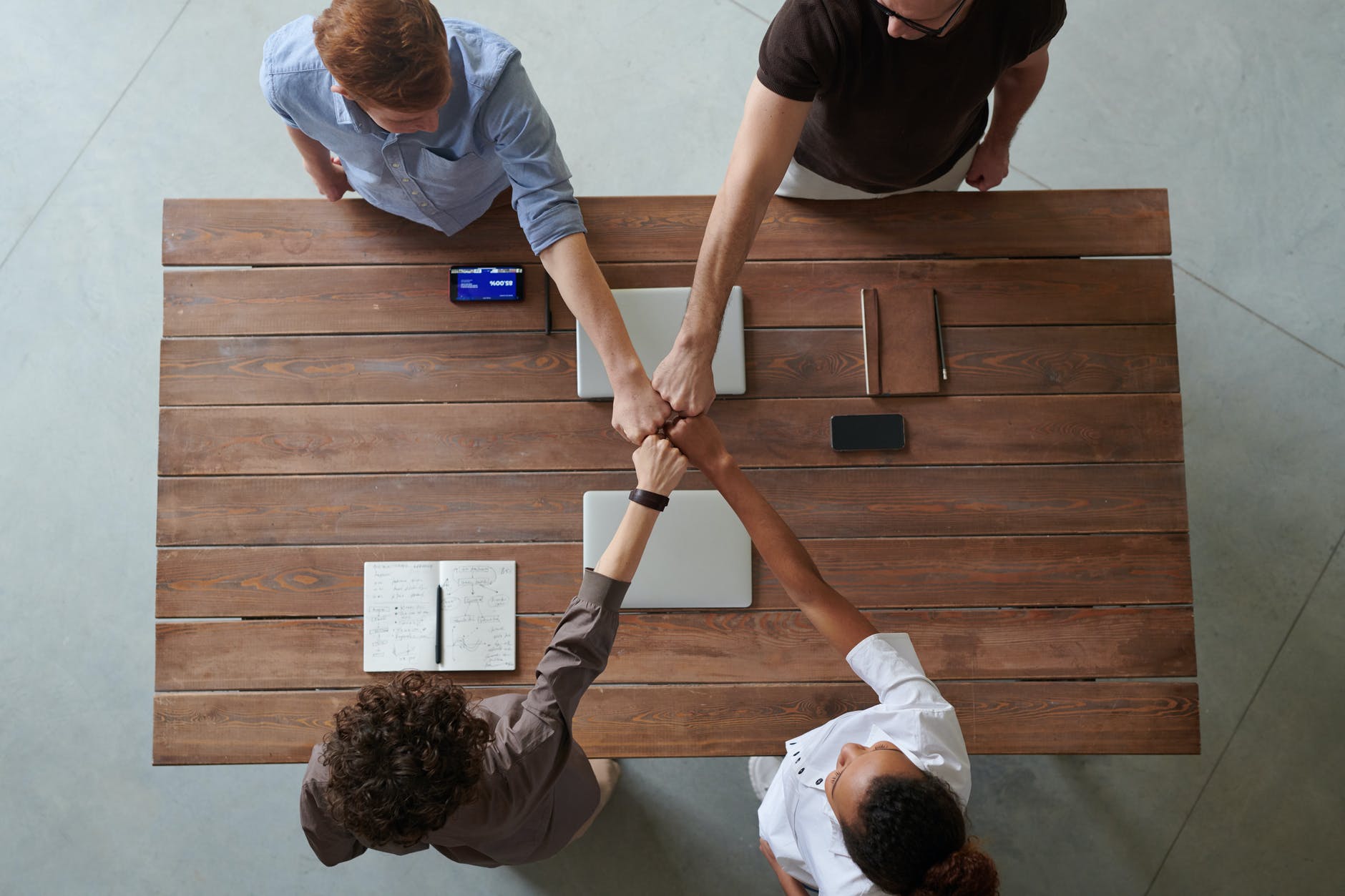 Top view of wooden table and four people around it, each with one hand in fist extended connecting all at the center. Different skin tones are visible.