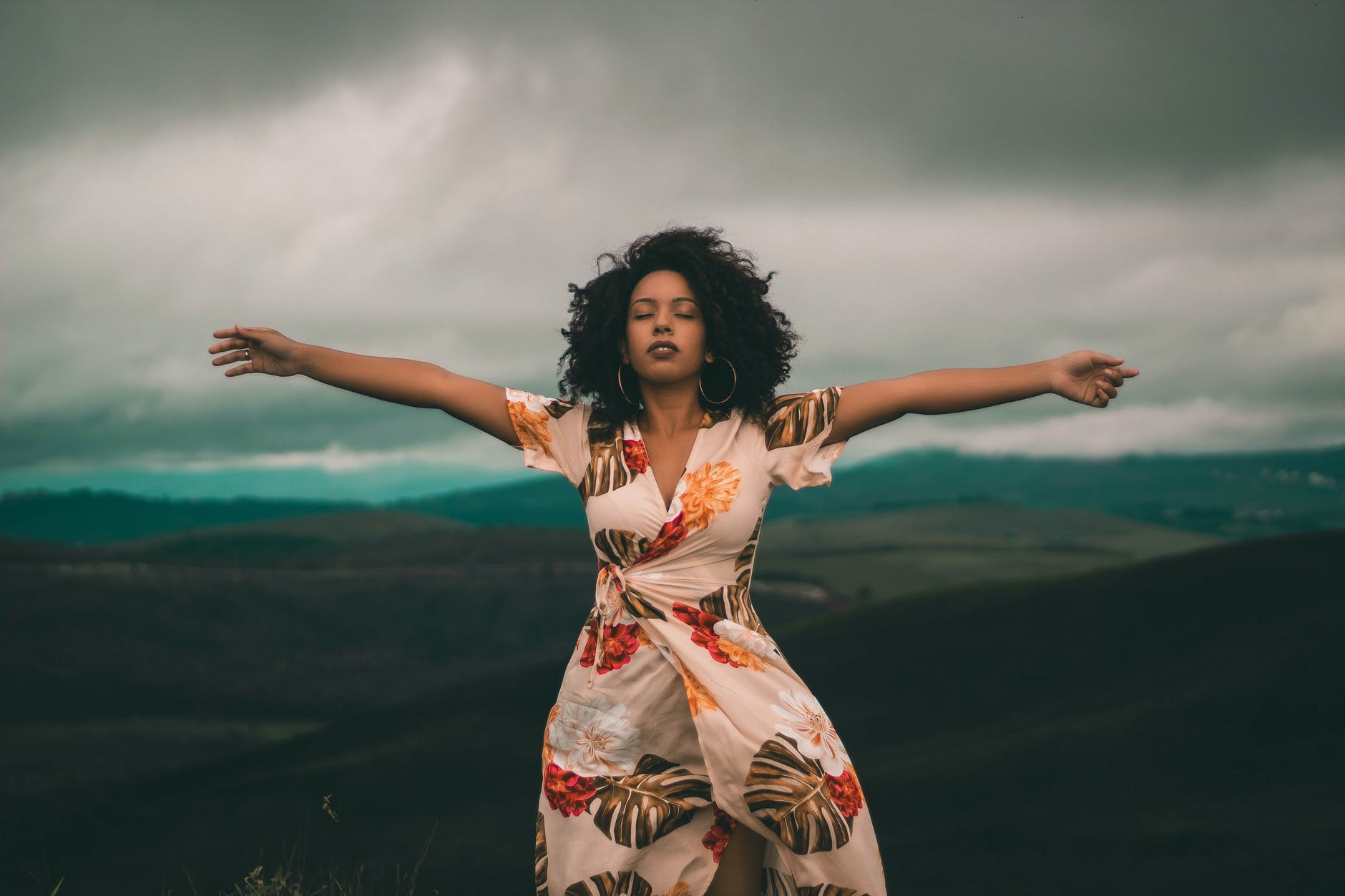 Black woman with curly black hair with arms spread out and eyes closed. The woman is wearing a white dress with flower/plant pattern. The background is cloudy with mountains.