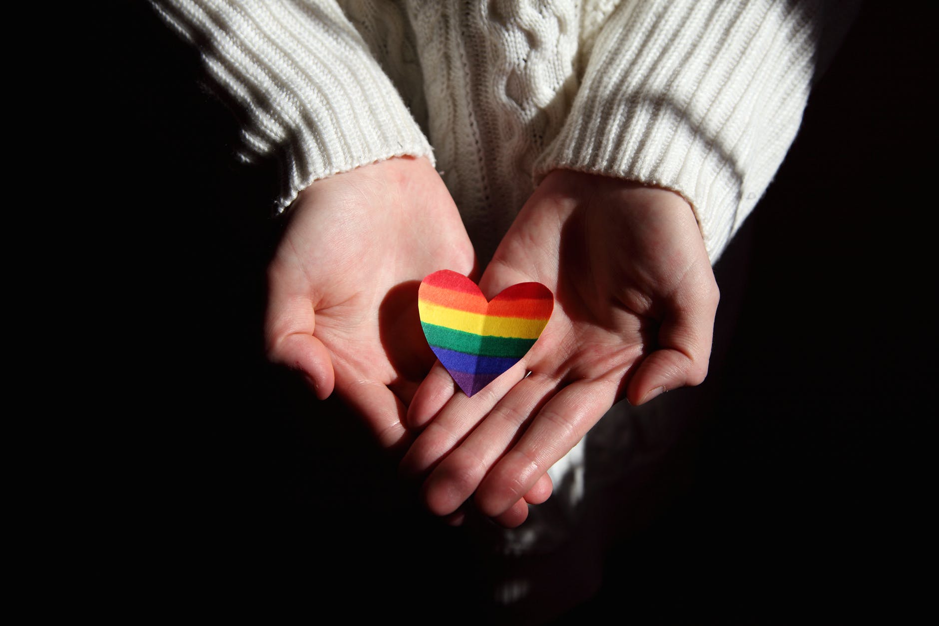 Pair of hands holding rainbow colored heart. Hands do not have jewelry and person is wearing white sweater.