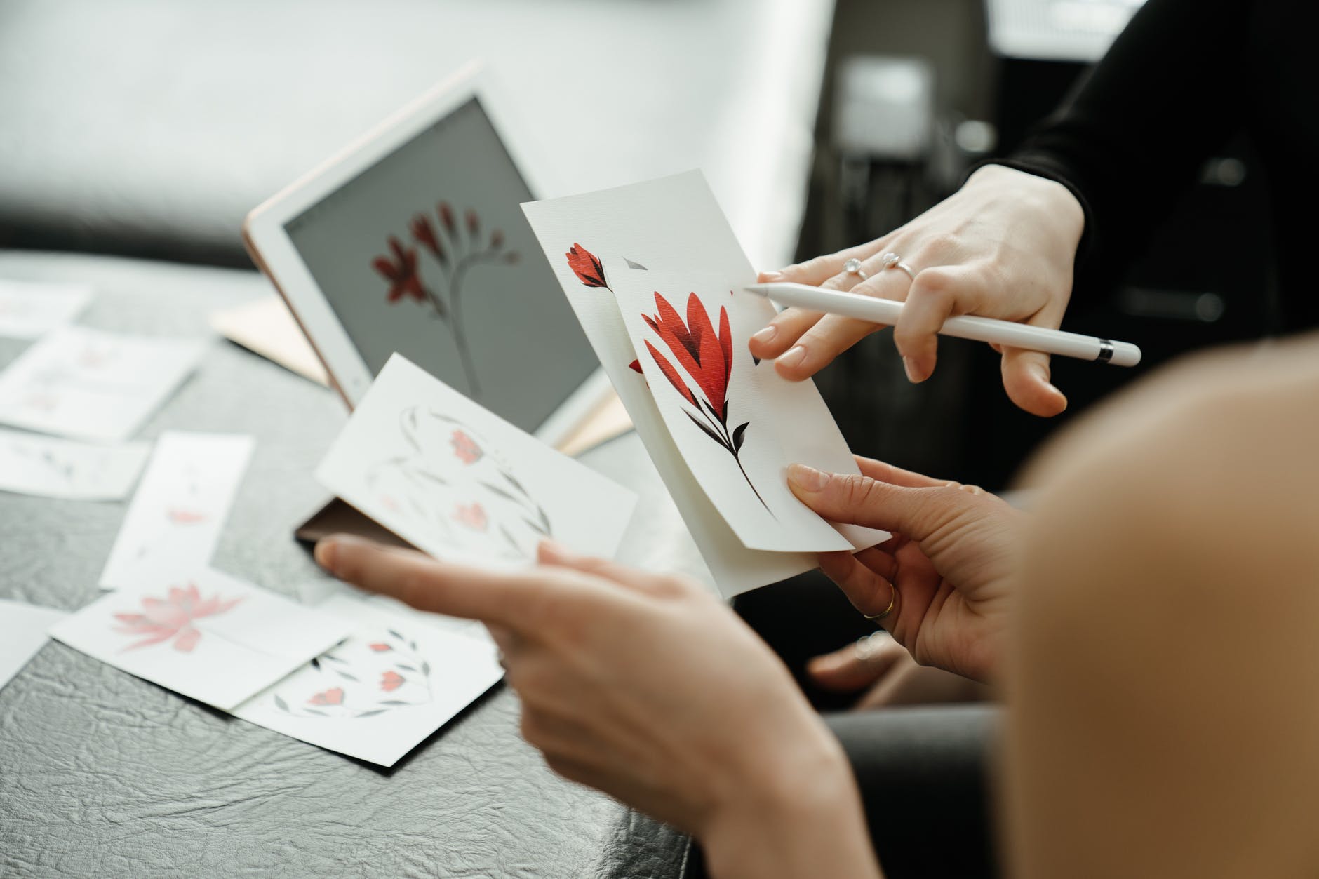 Hands holding drawings of red flowers. A hand holding a white stylus can be seen pointing to one drawing. A tablet is visible in the background.