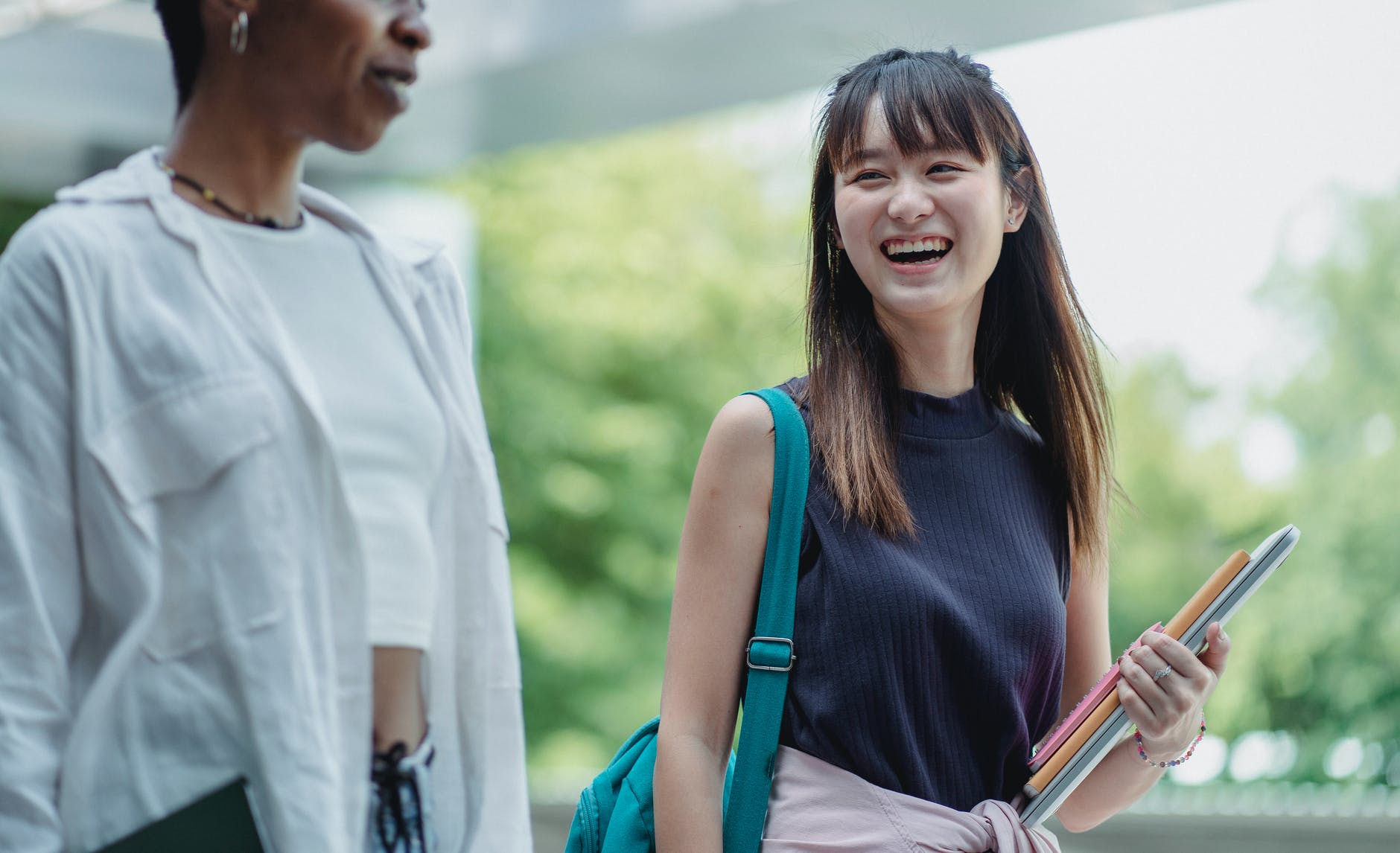 Asian student with long hair and bangs holding books in left hand and smiling walking with black woman.