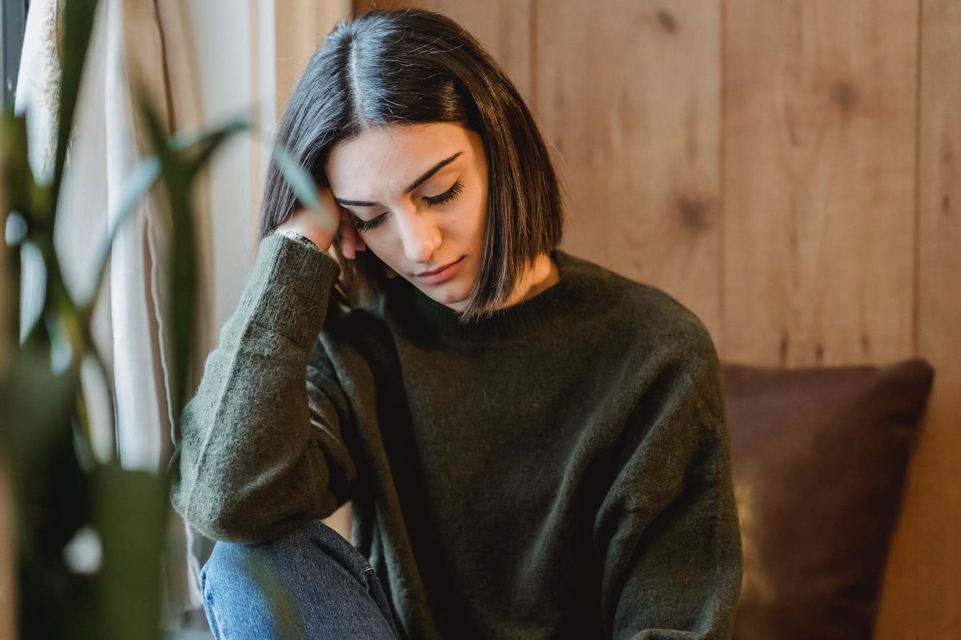 Woman with shoulder-length brown hair and dark green sweater looking down resting head on hand near cheek.