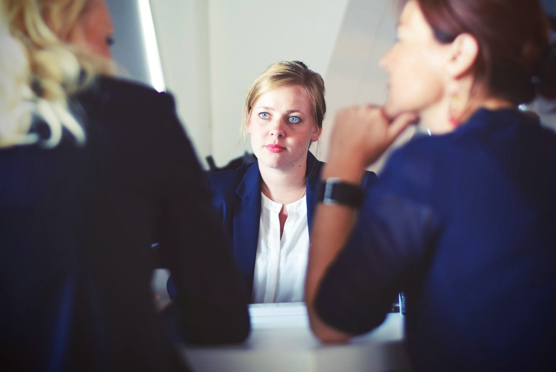 Woman with blond hair and blue eyes seated facing two people that are out of focus. One is facing the other one.