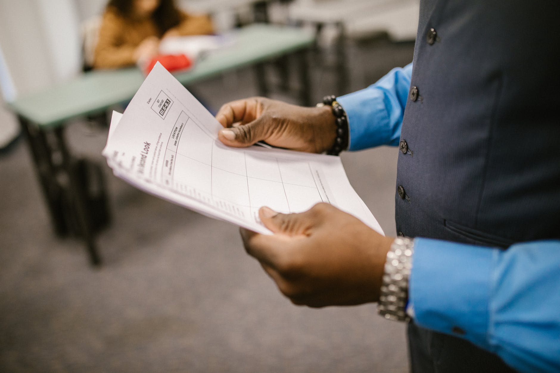 Person with dark skin holding paper with tables. Hands are visible and a vest. Tables/desks are blurred in the background.