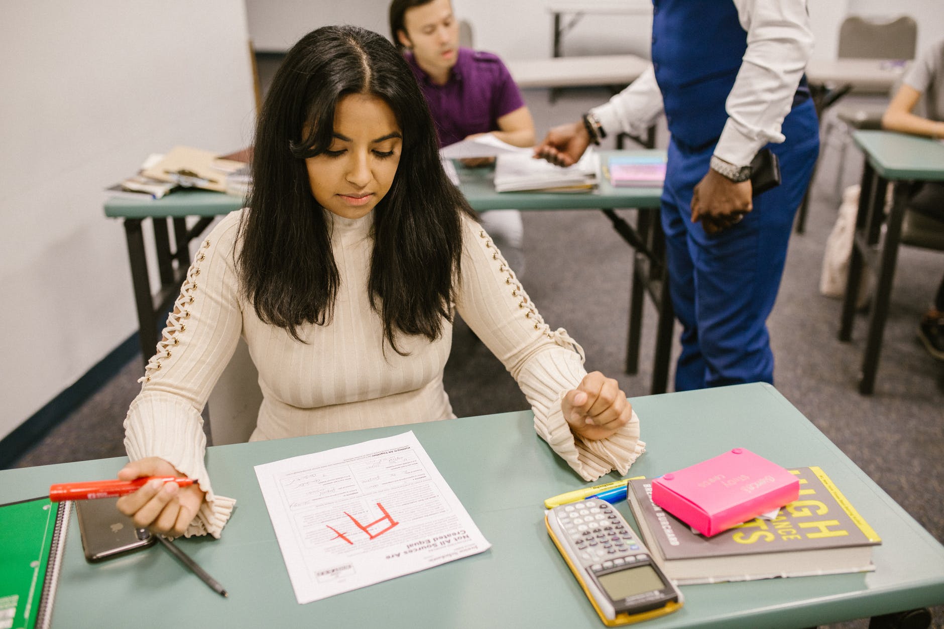 Student at desk staring at exam with A+ in red marker. Student is holding red marker in right hand. The desk has a calculator, highlighters, and a book.