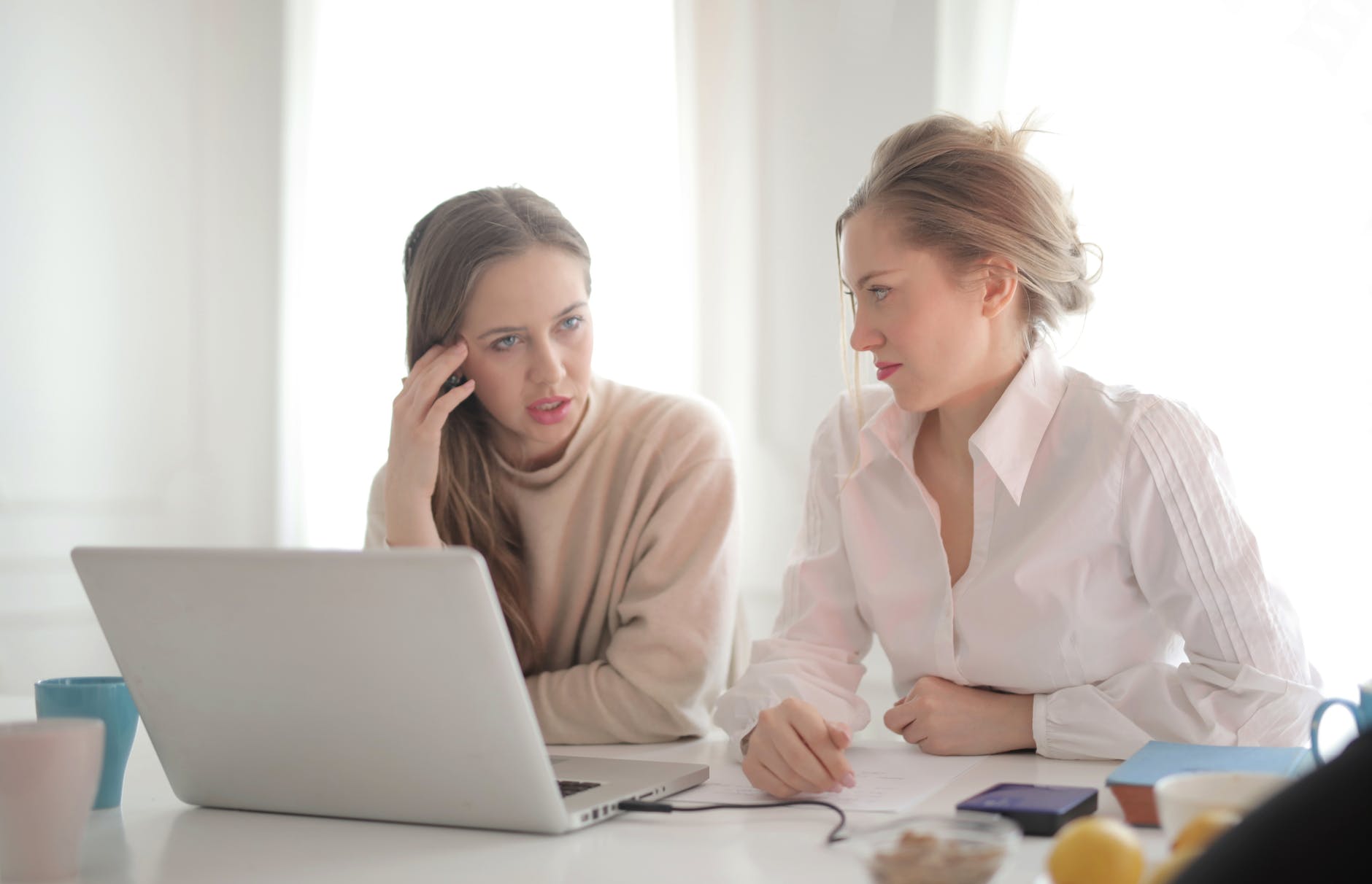 Two women seated in front of laptop. One is looking at the other one who is deep in thought.