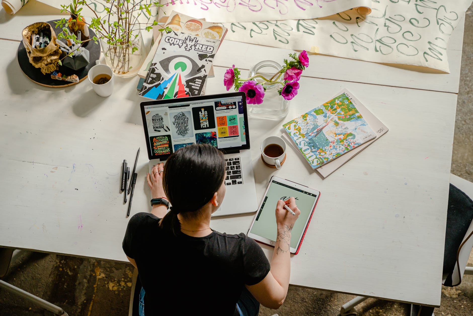Woman writing on tablet with right hand and using laptop. Desk has coffee and magazines and flowers.