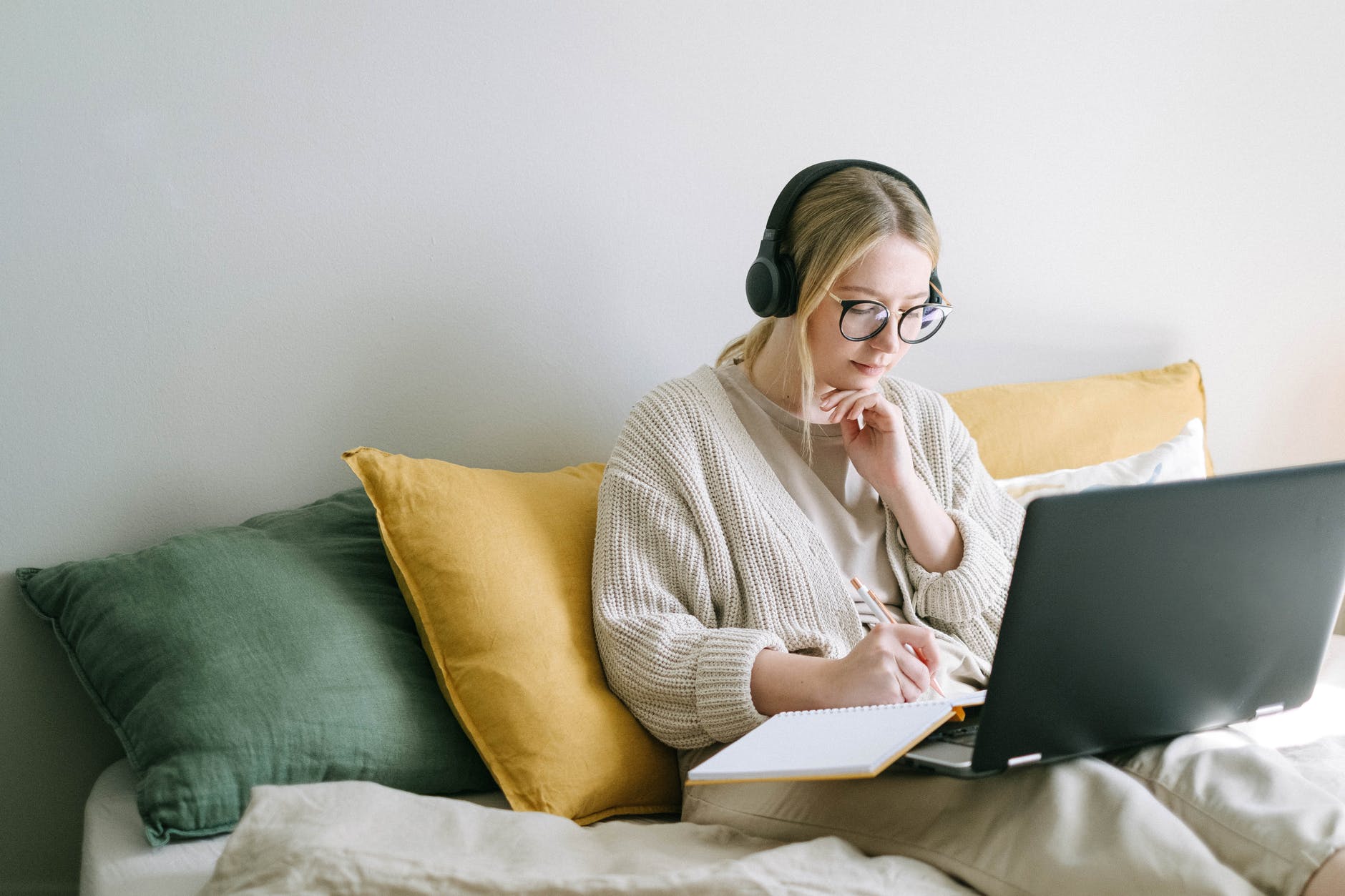 Woman seated on couch with yellow cushions with large black laptop and note book.