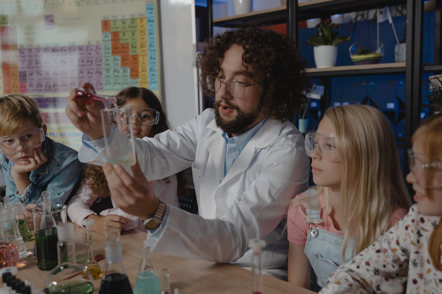 Instructor wearing white lab coat and holding beaker. Instructor is male with beard, long hair, and glasses. Two children wearing safety glasses are watching on either side. The table has several beakers and substances.