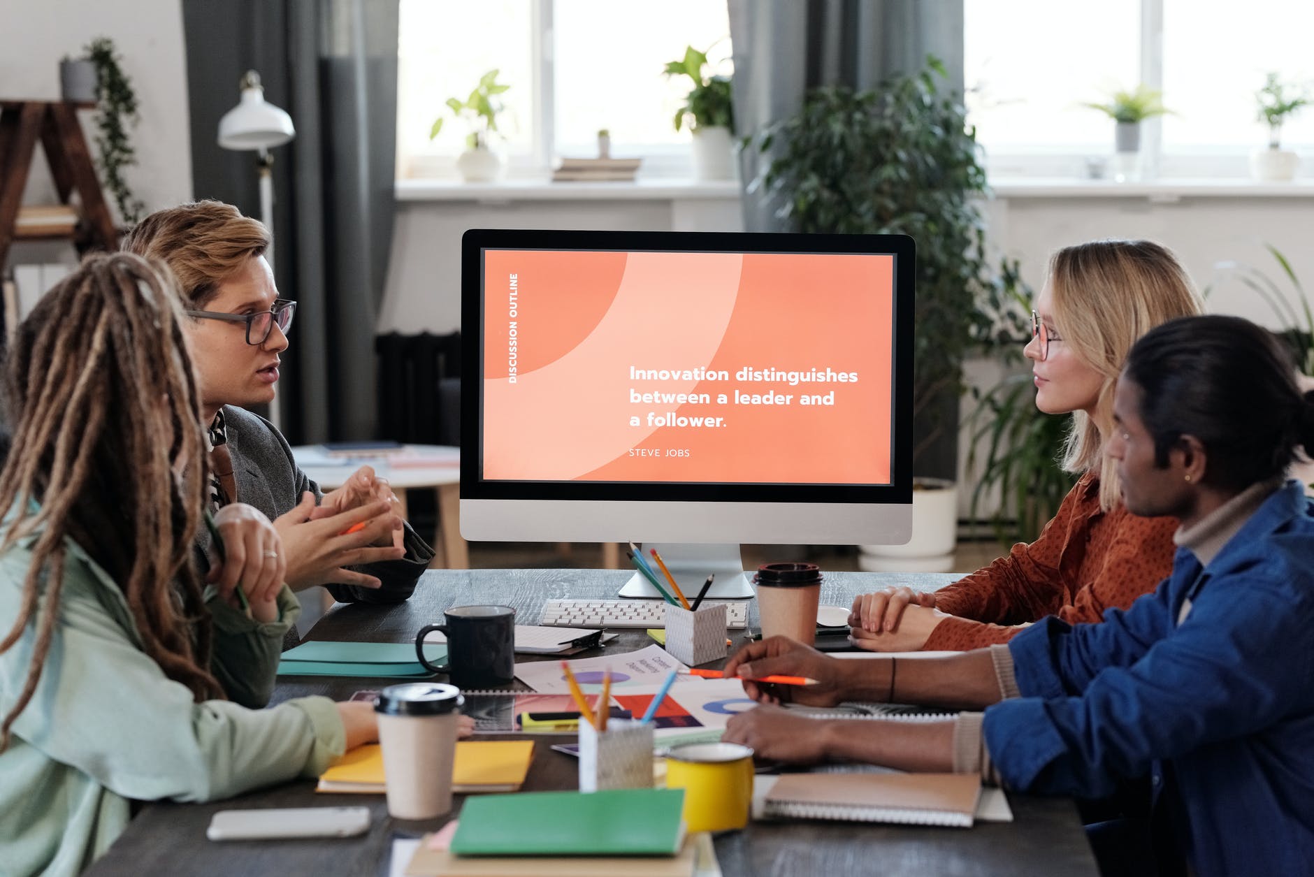 Group of people seated at table with notebooks and coffee cups. Two people on each side and computer monitor with "Innovation distinguishes between a leader and a follower. Steve Jobs" on the screen.